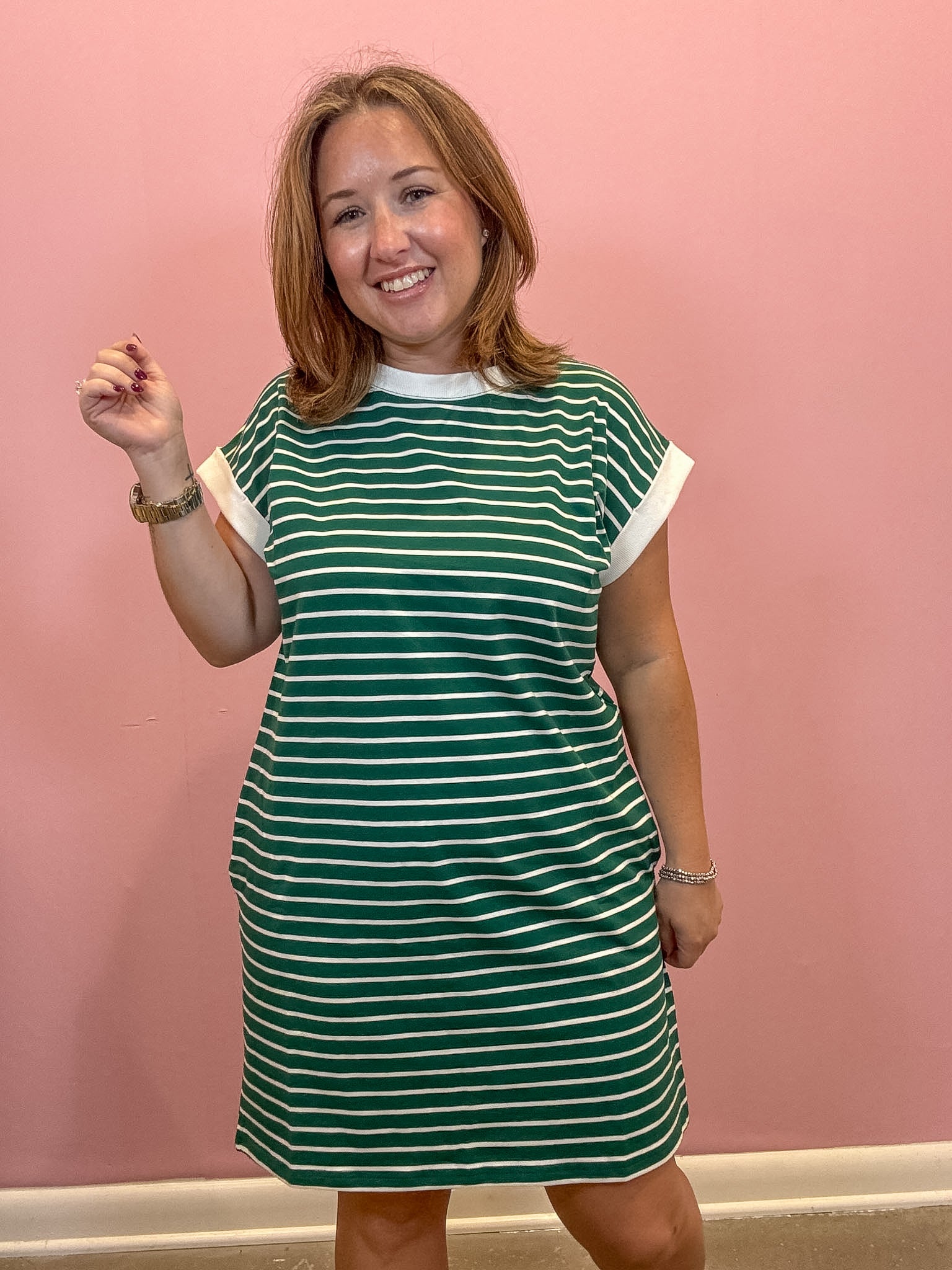 A woman modeling a green and white horizontally striped short-sleeve t-shirt dress against a solid pink wall.