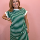 A woman modeling a green and white striped short-sleeve dress with solid white trim at the crew neck and sleeve cuffs, standing against a solid pink wall.