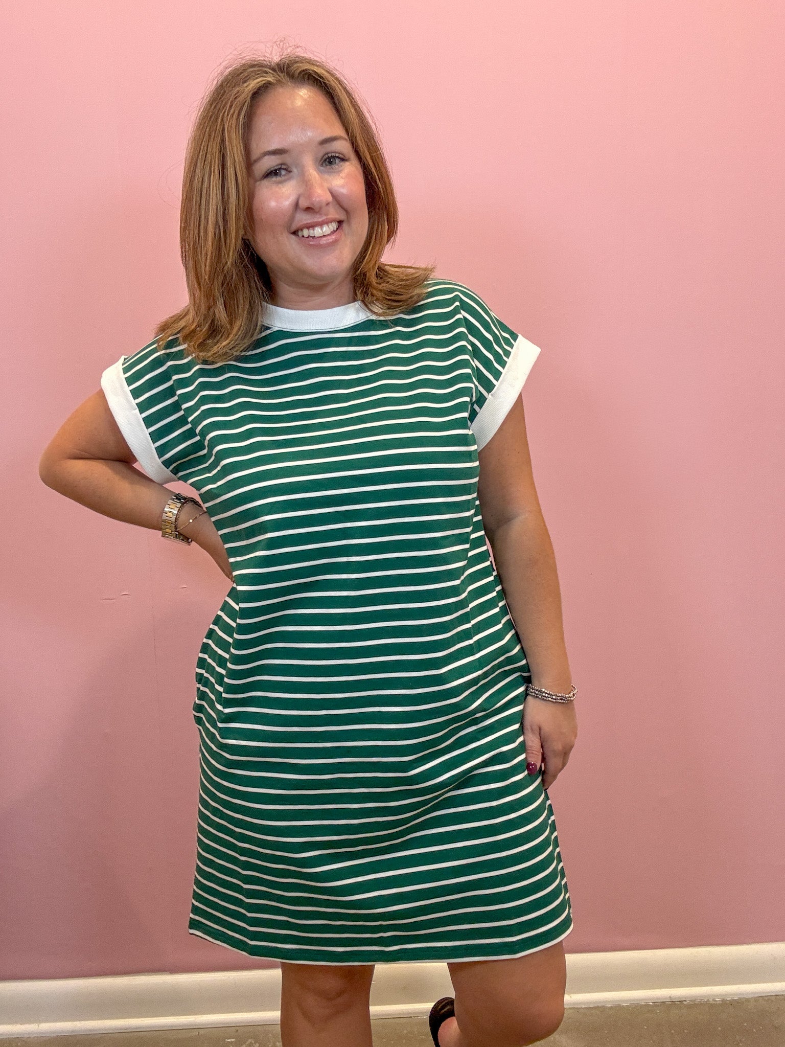 A woman modeling a green and white striped short-sleeve dress with solid white trim at the crew neck and sleeve cuffs, standing against a solid pink wall.
