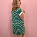 A woman wearing a green and white striped short-sleeve dress, standing against a solid pink wall.