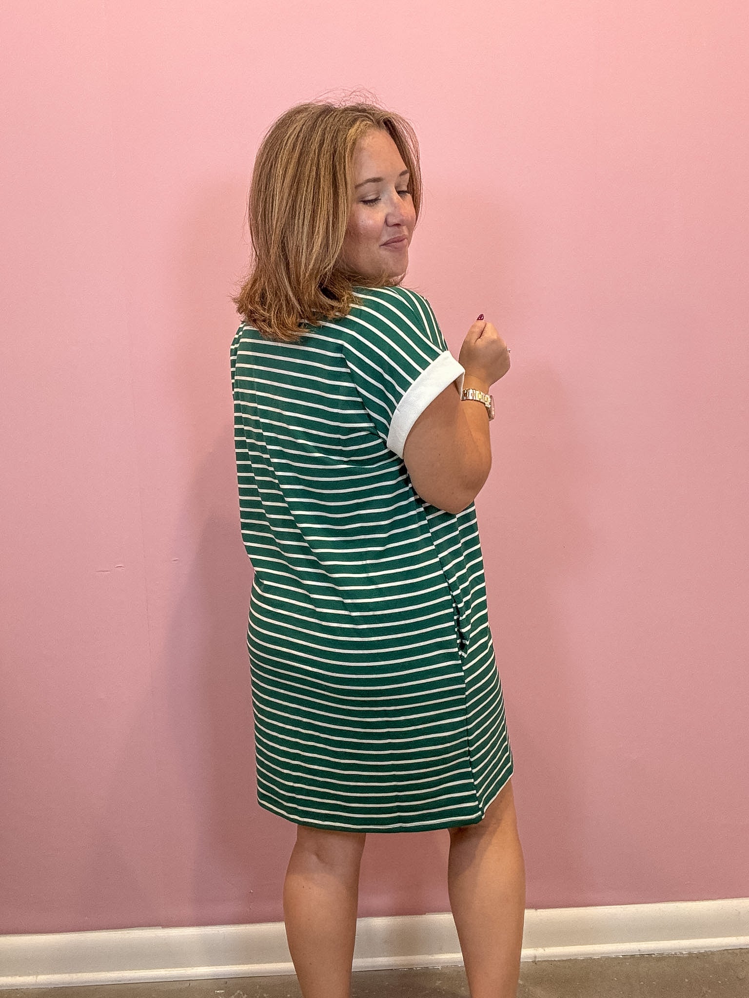A woman wearing a green and white striped short-sleeve dress, standing against a solid pink wall.