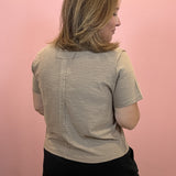 A woman with shoulder-length blonde hair is shown from the back, wearing a light gray short-sleeve textured top and black shorts against a solid pink wall.