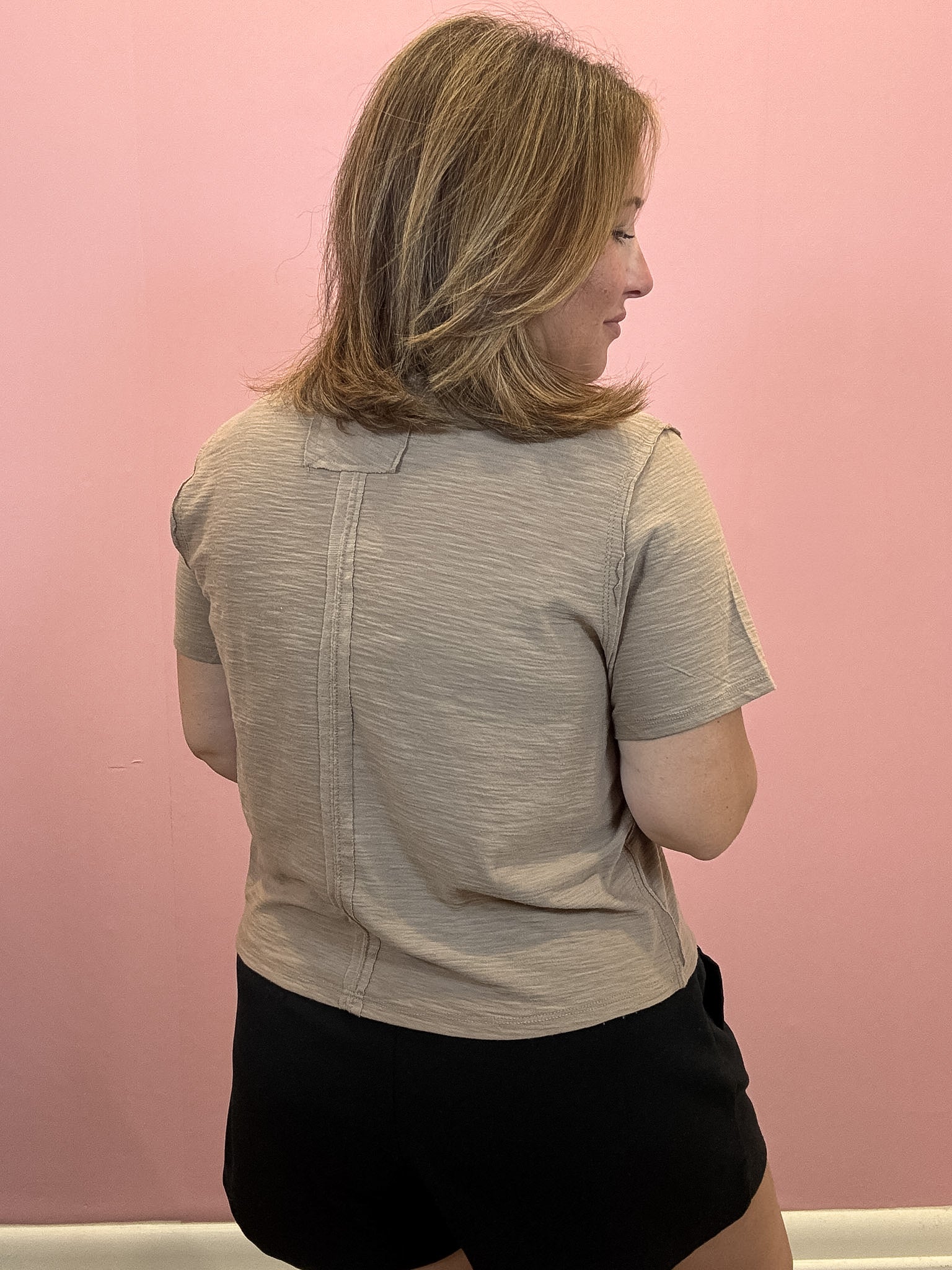 A woman with shoulder-length blonde hair is shown from the back, wearing a light gray short-sleeve textured top and black shorts against a solid pink wall.