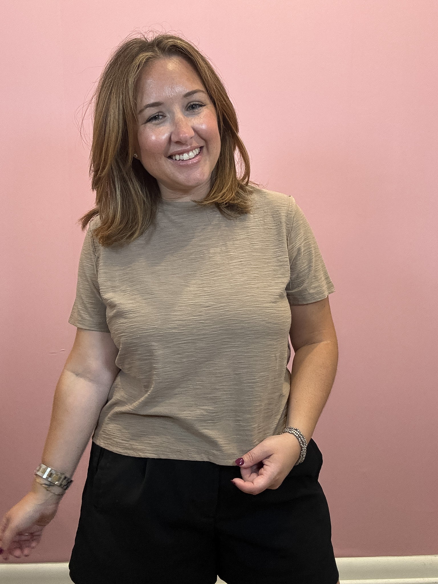 A smiling woman with shoulder-length light brown hair wearing a beige short-sleeve crewneck t-shirt and black pants, standing against a solid pink wall.