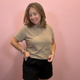 A woman modeling a short-sleeve crewneck t-shirt in a heathered beige color, paired with black shorts, standing against a solid pink wall.