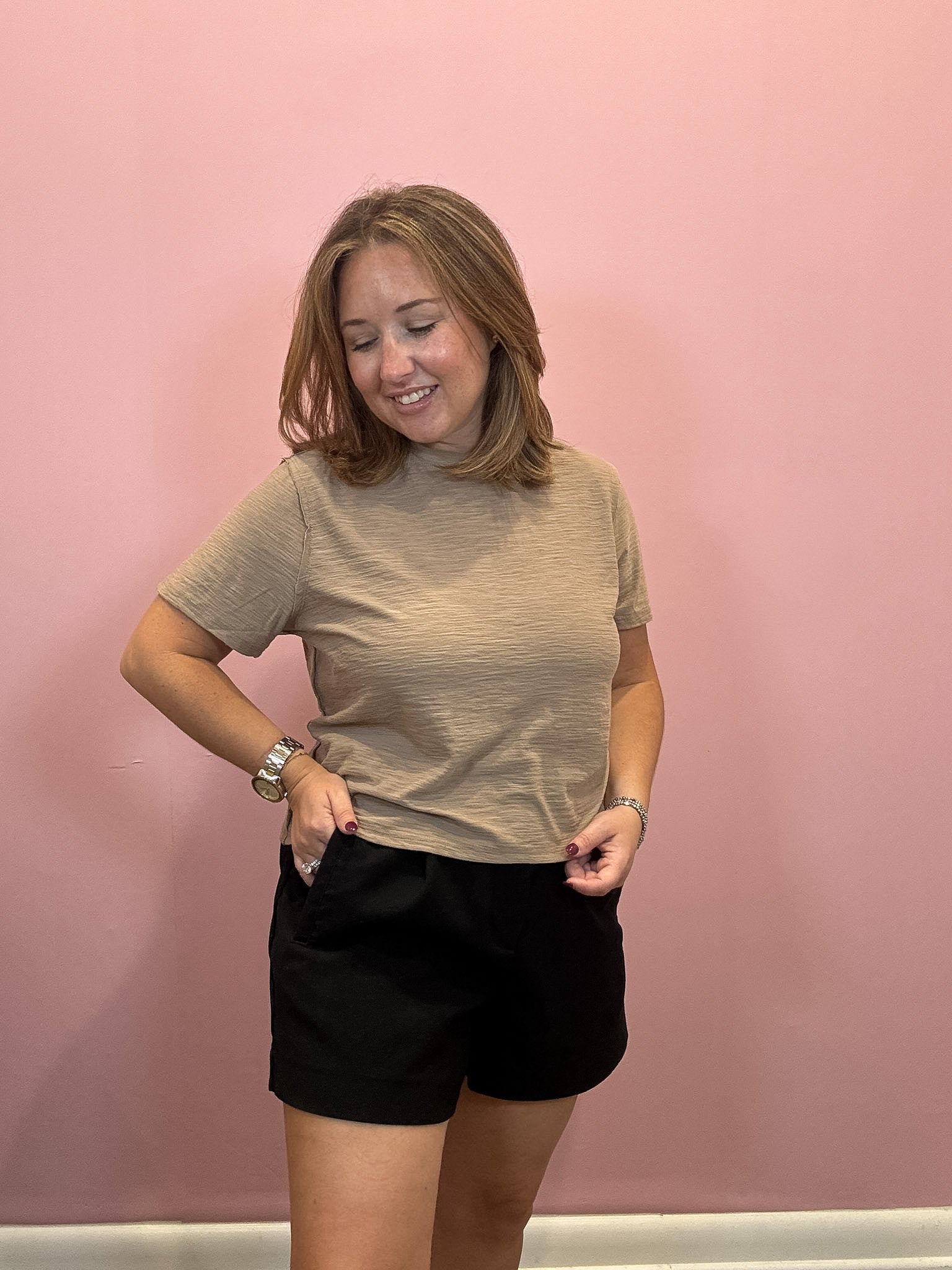 A woman modeling a short-sleeve crewneck t-shirt in a heathered beige color, paired with black shorts, standing against a solid pink wall.