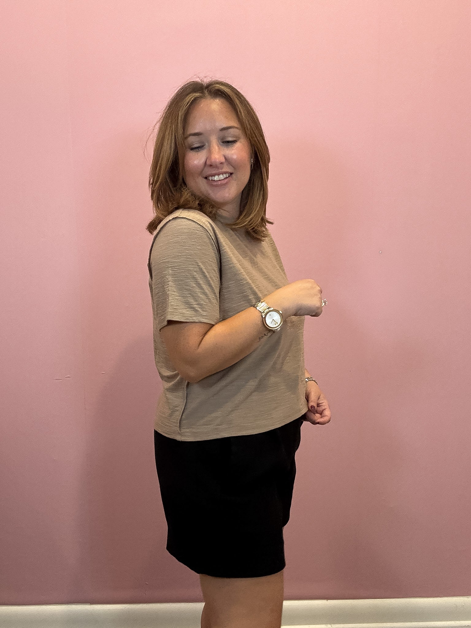 A woman modeling a beige short-sleeve t-shirt and black skirt against a solid pink wall.