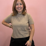 A woman with shoulder-length brown hair smiling in front of a pink wall, wearing a beige short-sleeve crewneck t-shirt and black shorts.