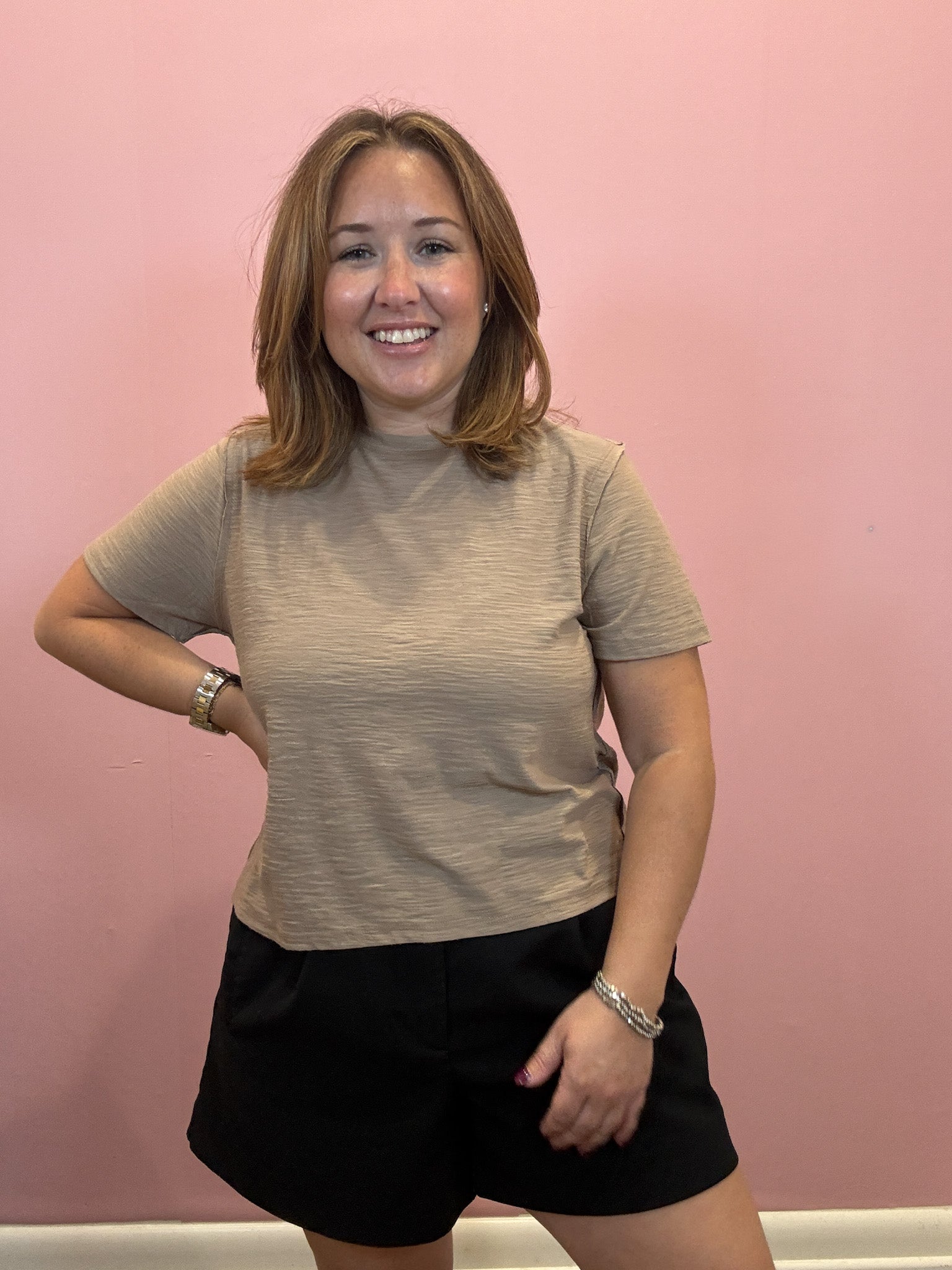 A woman with shoulder-length brown hair smiling in front of a pink wall, wearing a beige short-sleeve crewneck t-shirt and black shorts.