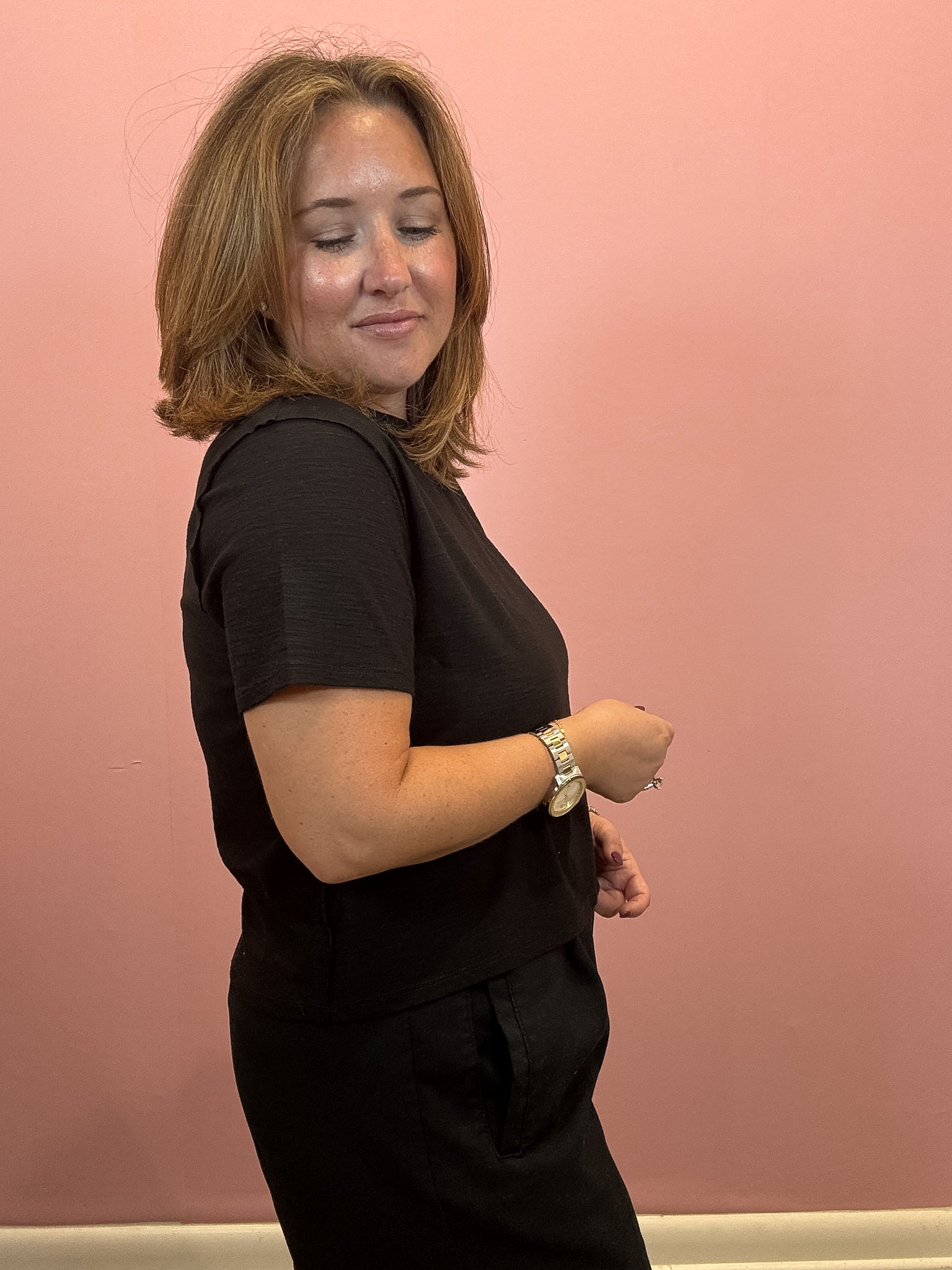 A woman with shoulder-length light brown hair is shown in profile wearing a black short-sleeve top and black bottoms, standing against a solid pink wall.