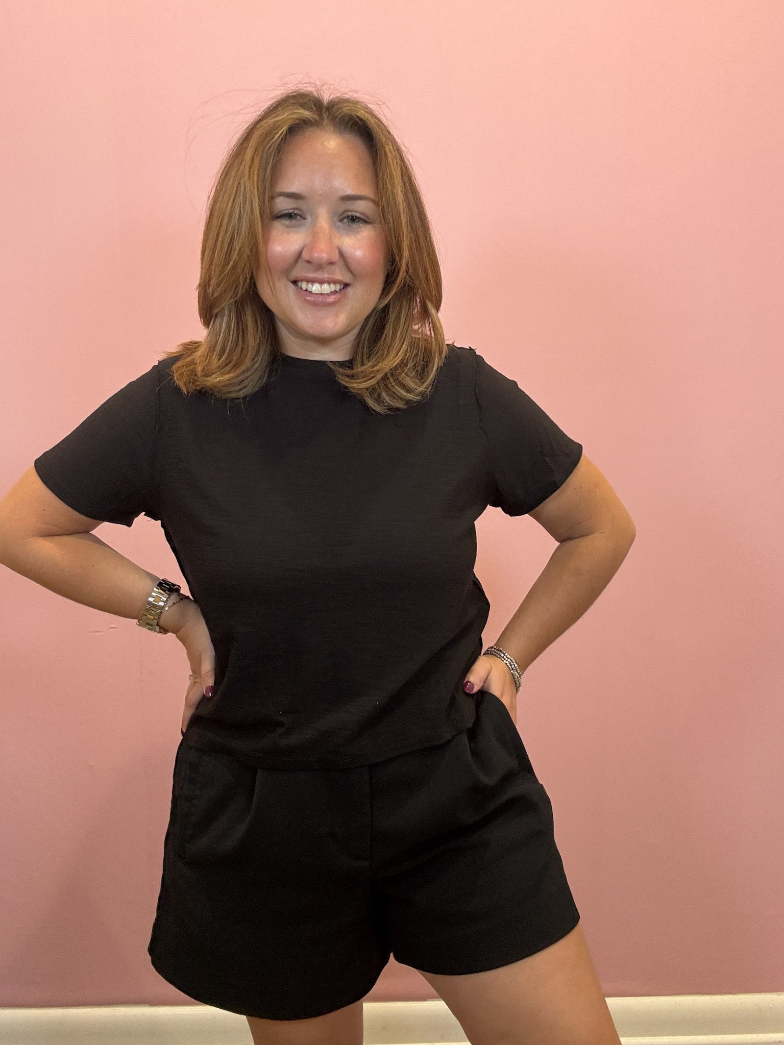 A woman with shoulder-length light brown hair smiling, wearing a black short-sleeve crewneck t-shirt and black shorts, posing with hands on hips against a solid pink wall.