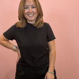 A woman with shoulder-length light brown hair smiling, wearing a black short-sleeve t-shirt and black shorts against a solid pink wall.
