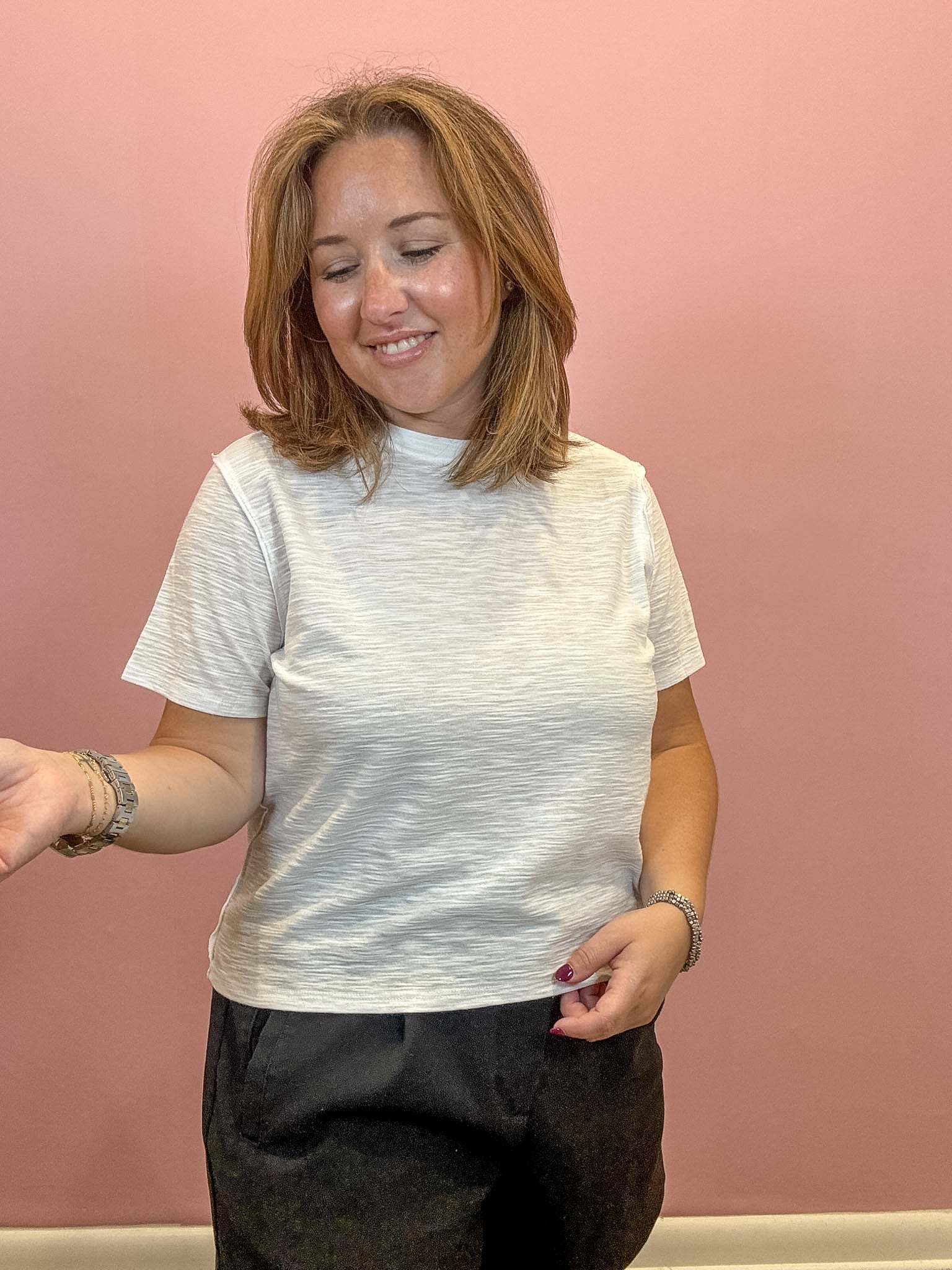 A woman with shoulder-length light brown hair is wearing a short-sleeve crewneck t-shirt in a heathered off-white color, paired with dark gray pants, standing against a solid dusty pink wall.
