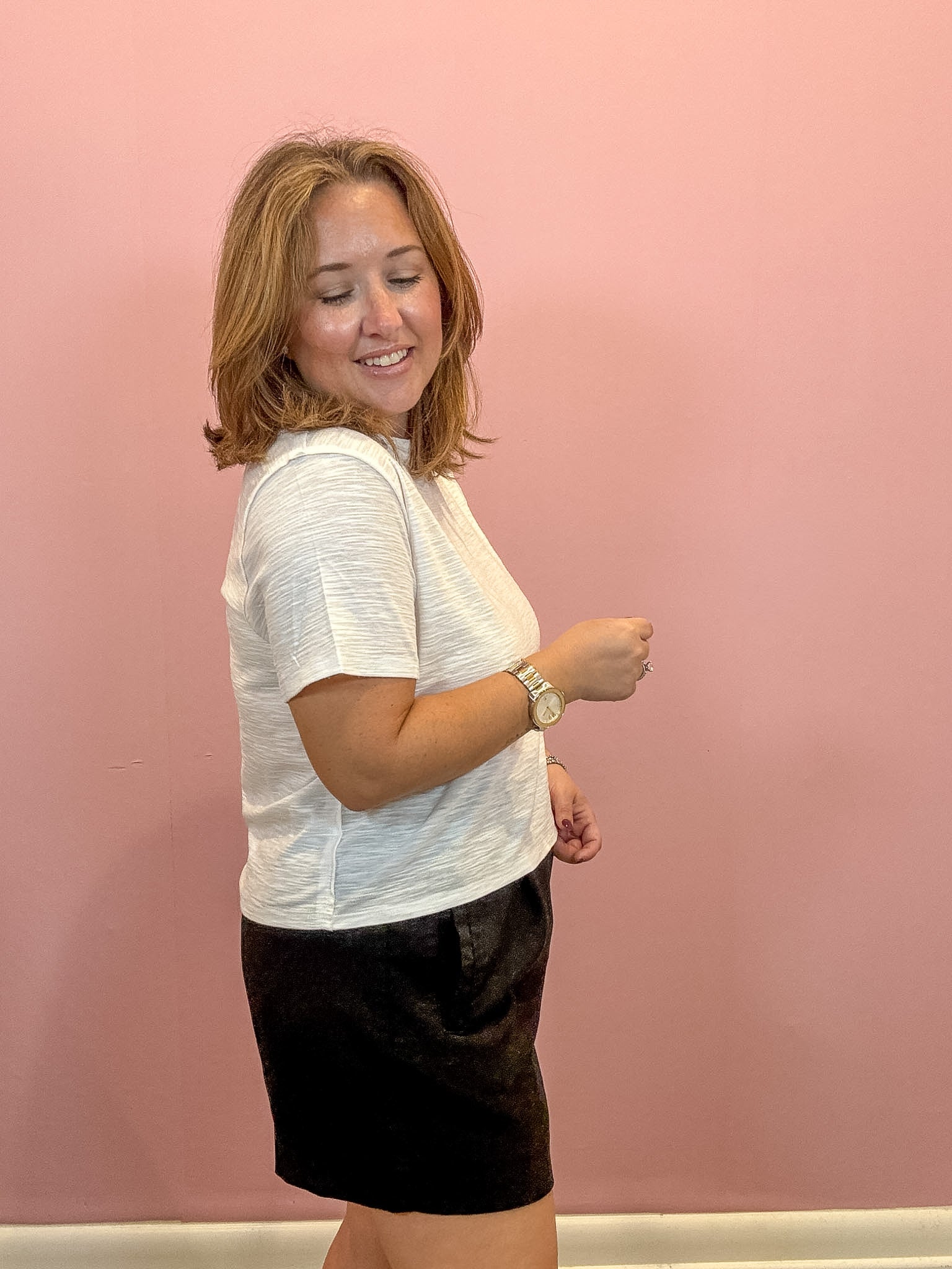 A woman with shoulder-length blonde hair is shown from the side, wearing a white textured short-sleeve t-shirt and a black skirt, standing against a solid pink wall.
