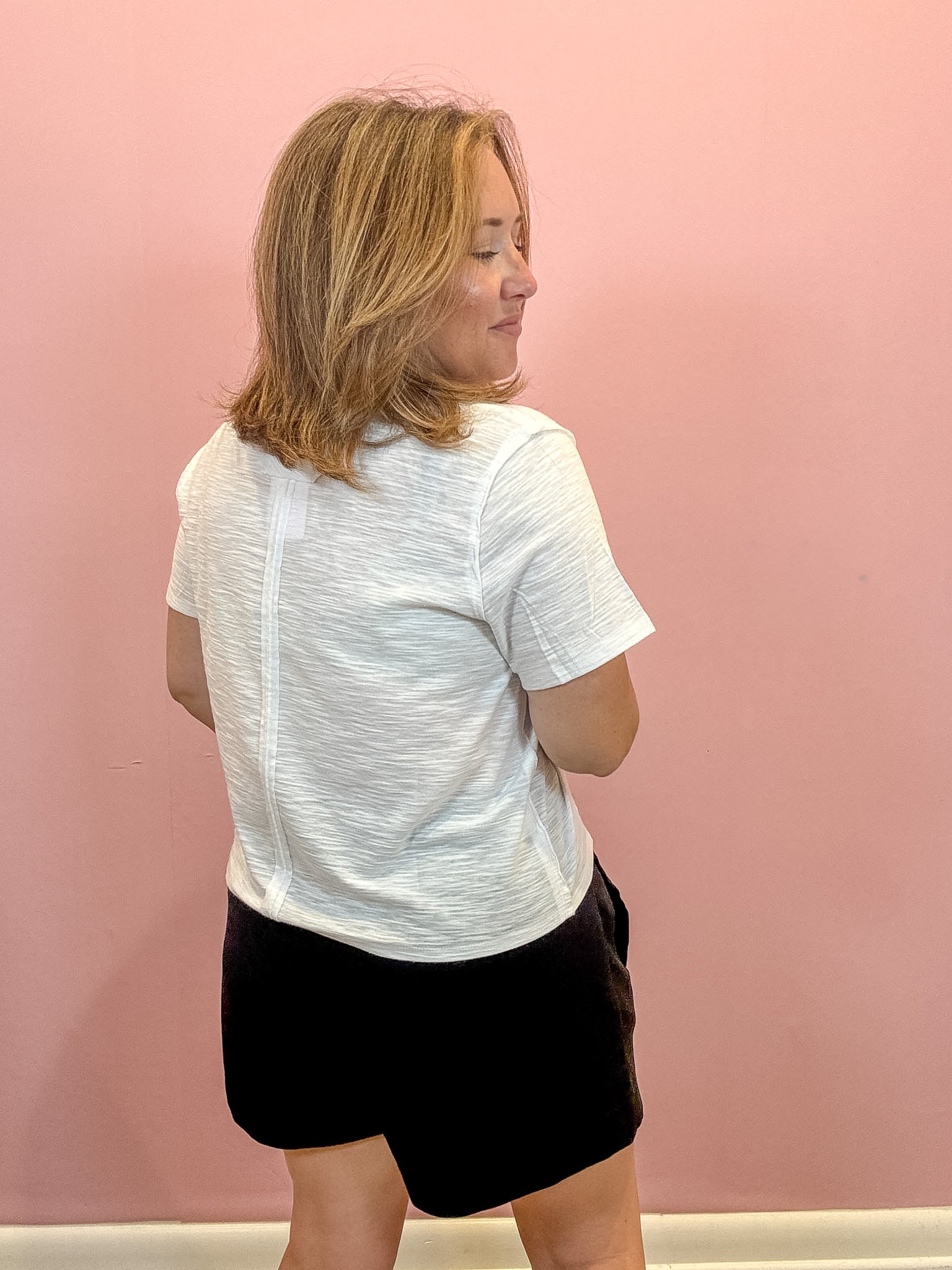 A woman with blonde hair is shown from the back wearing a white heathered short-sleeve t-shirt and black shorts, standing against a solid pink wall.