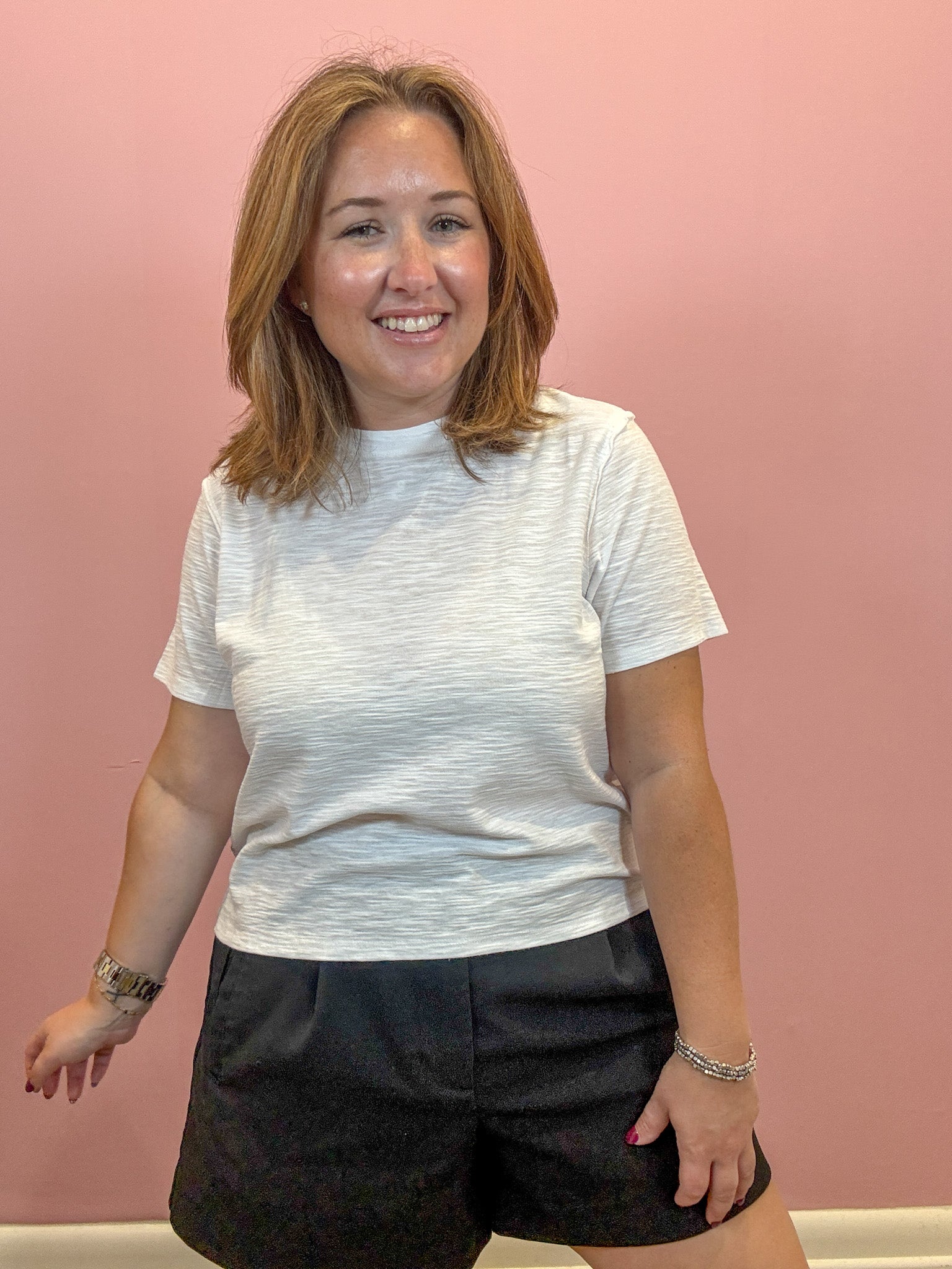 A woman with shoulder-length light brown hair smiling, wearing a white short-sleeve crewneck t-shirt and black shorts, standing against a solid pink wall.