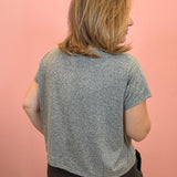 A woman with shoulder-length blonde hair is shown from behind wearing a short-sleeve heather gray t-shirt and dark shorts, standing against a solid pink wall.