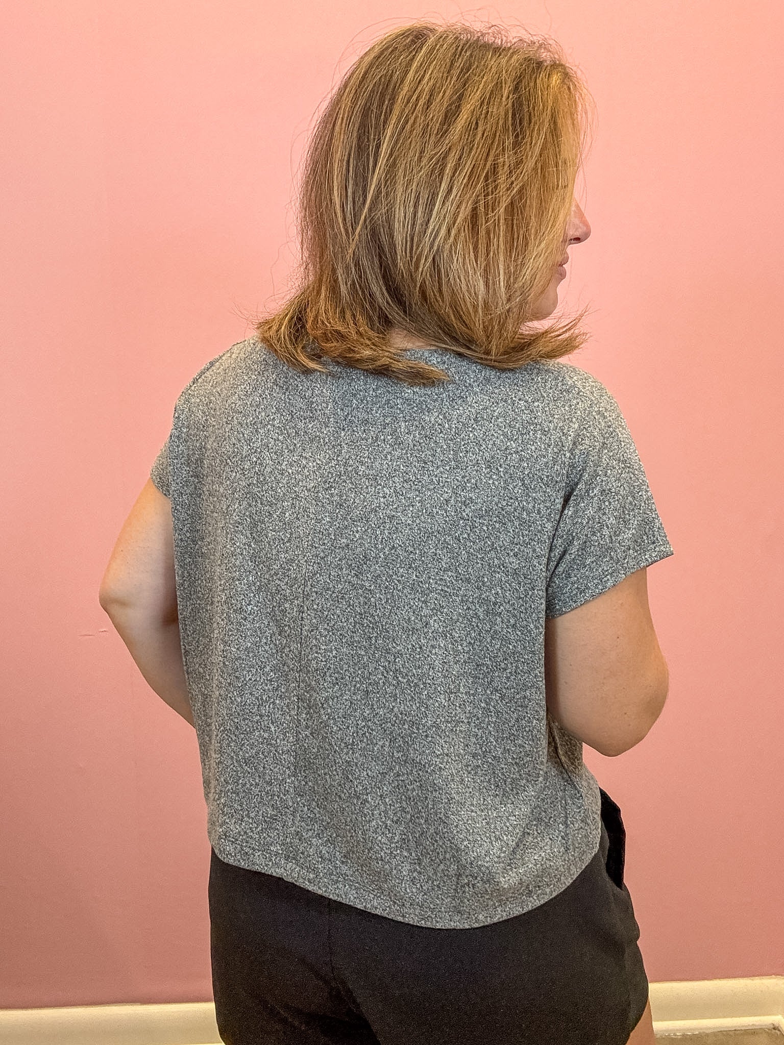 A woman with shoulder-length blonde hair is shown from behind wearing a short-sleeve heather gray t-shirt and dark shorts, standing against a solid pink wall.
