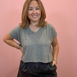 A woman with shoulder-length light brown hair stands against a solid pink wall, wearing a short-sleeve gray knit top and dark shorts.