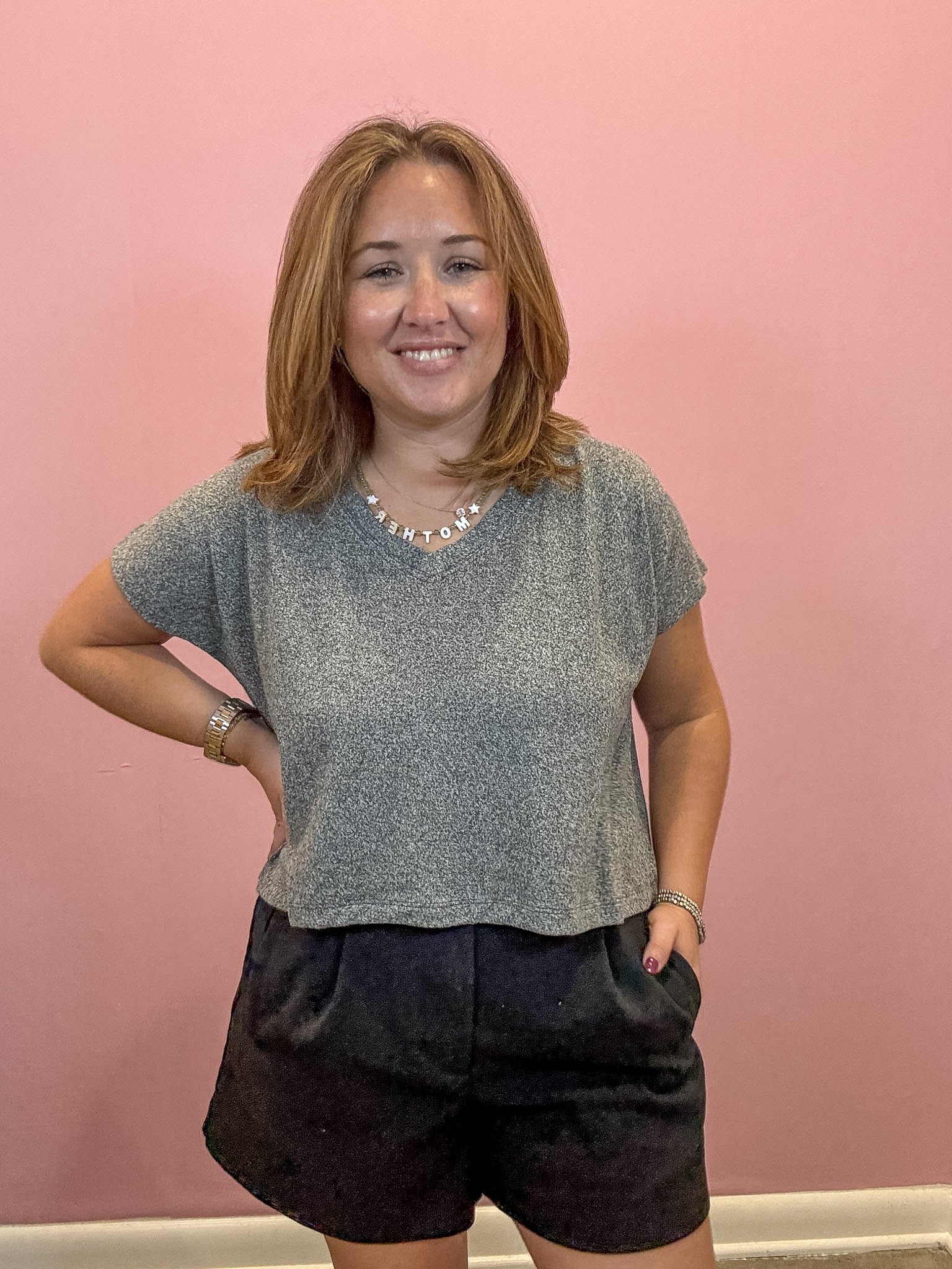 A woman with shoulder-length light brown hair stands against a solid pink wall, wearing a short-sleeve gray knit top and dark shorts.