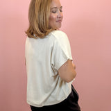 A woman with shoulder-length blonde hair is shown from the back, wearing a loose-fitting off-white t-shirt and black shorts against a solid pink wall.