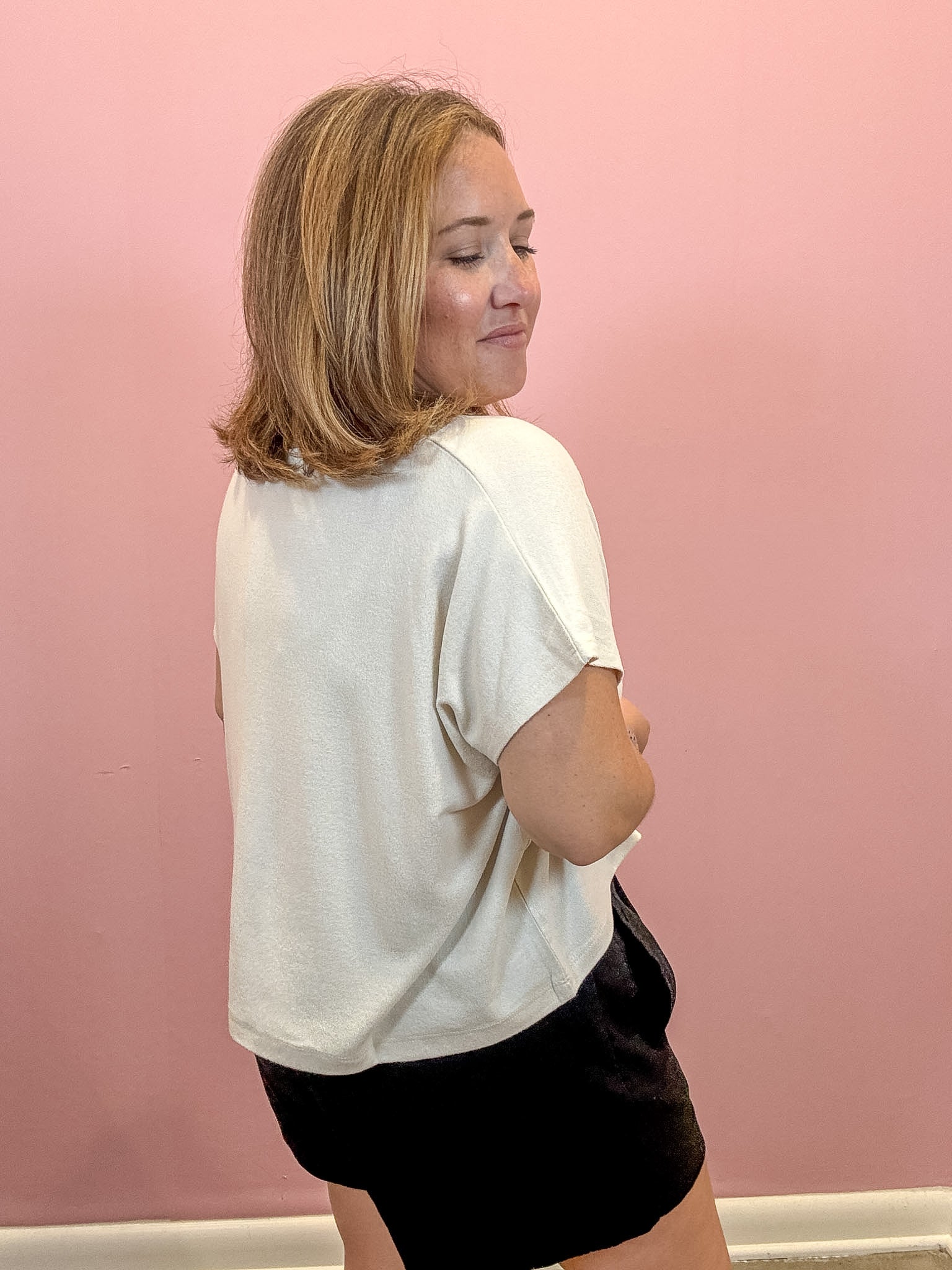 A woman with shoulder-length blonde hair is shown from the back, wearing a loose-fitting off-white t-shirt and black shorts against a solid pink wall.
