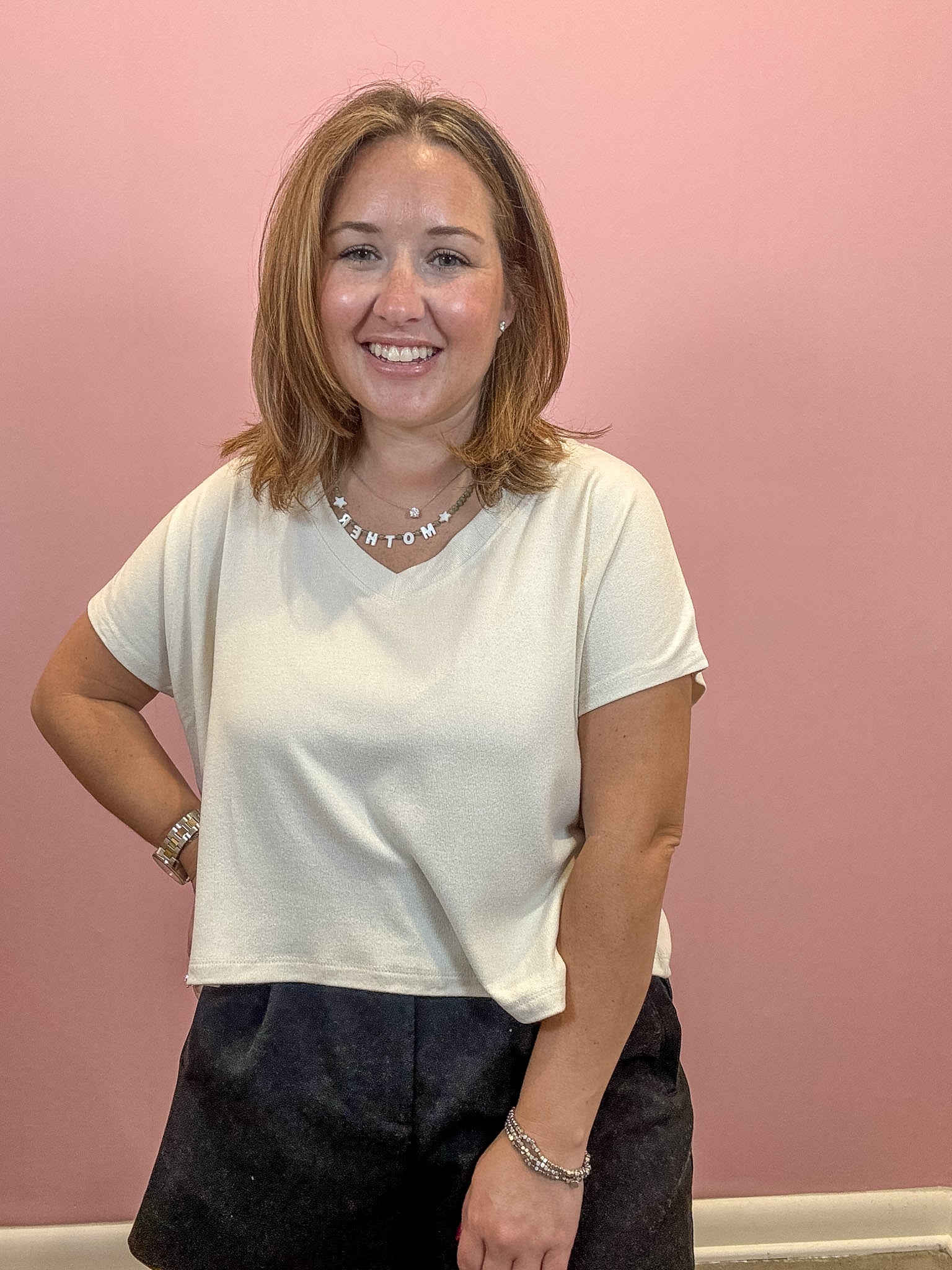 A smiling woman with shoulder-length brown hair stands against a solid pink wall, wearing a cream-colored short-sleeve V-neck top and dark charcoal shorts.