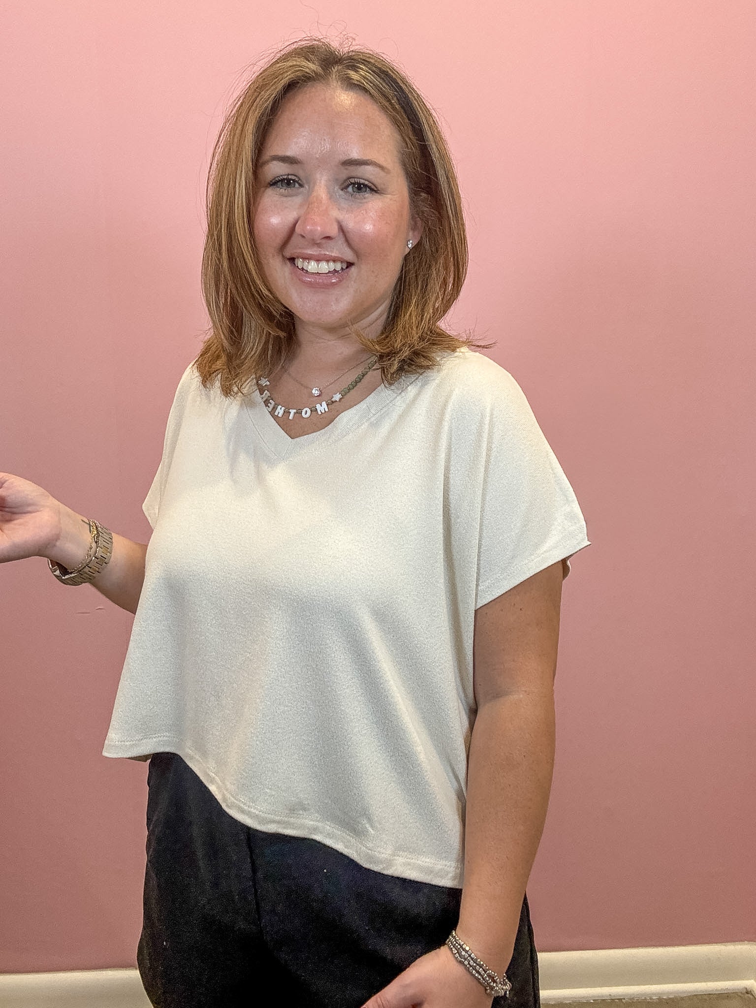 A woman with shoulder-length brown hair smiling, wearing a loose white short-sleeve V-neck top and dark pants, standing against a solid pink wall.
