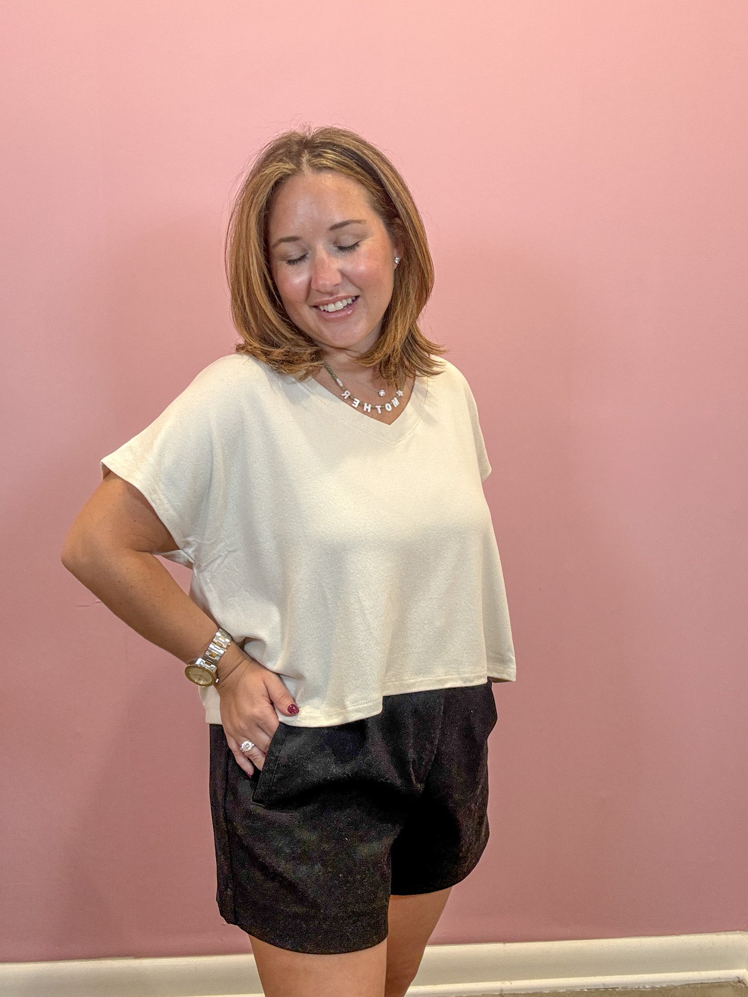 A woman posing in a cream-colored short-sleeve crop top and black shorts against a solid pink wall.