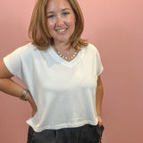 A smiling woman with shoulder-length light brown hair wearing a white short-sleeve V-neck t-shirt and dark gray pants, standing against a solid pink wall.