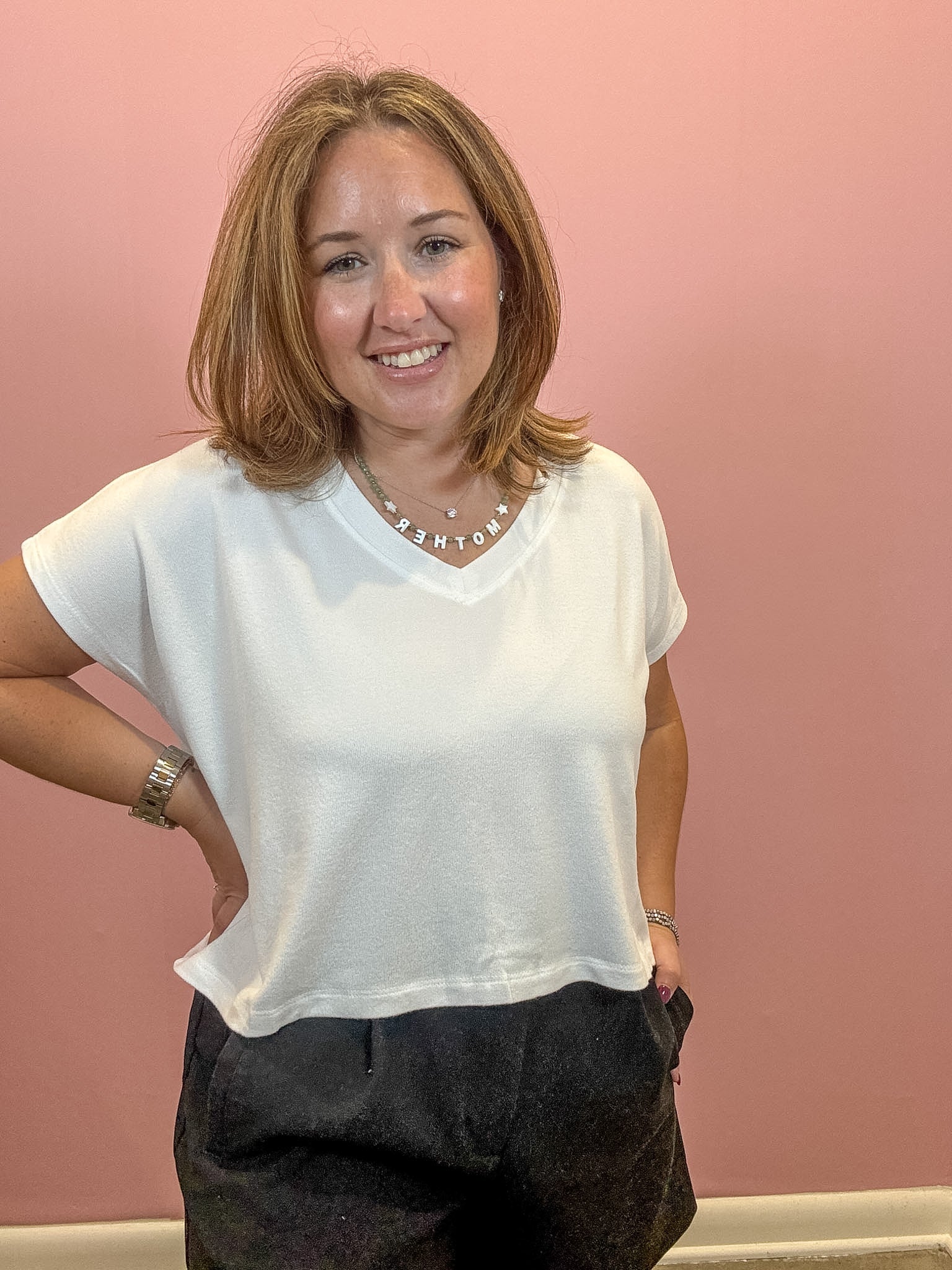 A smiling woman with shoulder-length light brown hair wearing a white short-sleeve V-neck t-shirt and dark gray pants, standing against a solid pink wall.