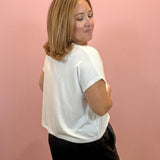 A woman with shoulder-length blonde hair is shown from the back, wearing a white short-sleeve t-shirt and black shorts, standing against a solid pink wall.