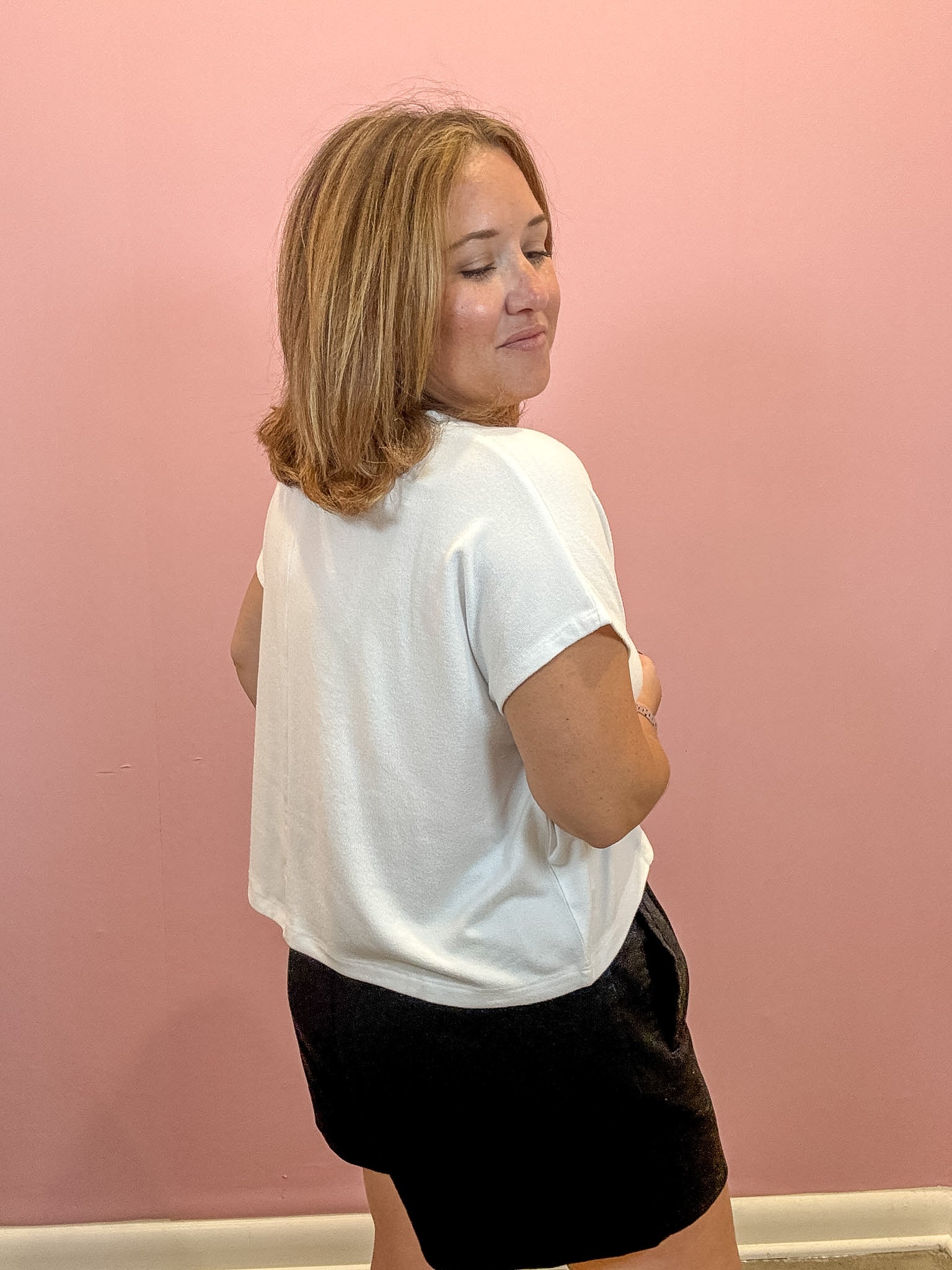 A woman with shoulder-length blonde hair is shown from the back, wearing a white short-sleeve t-shirt and black shorts, standing against a solid pink wall.