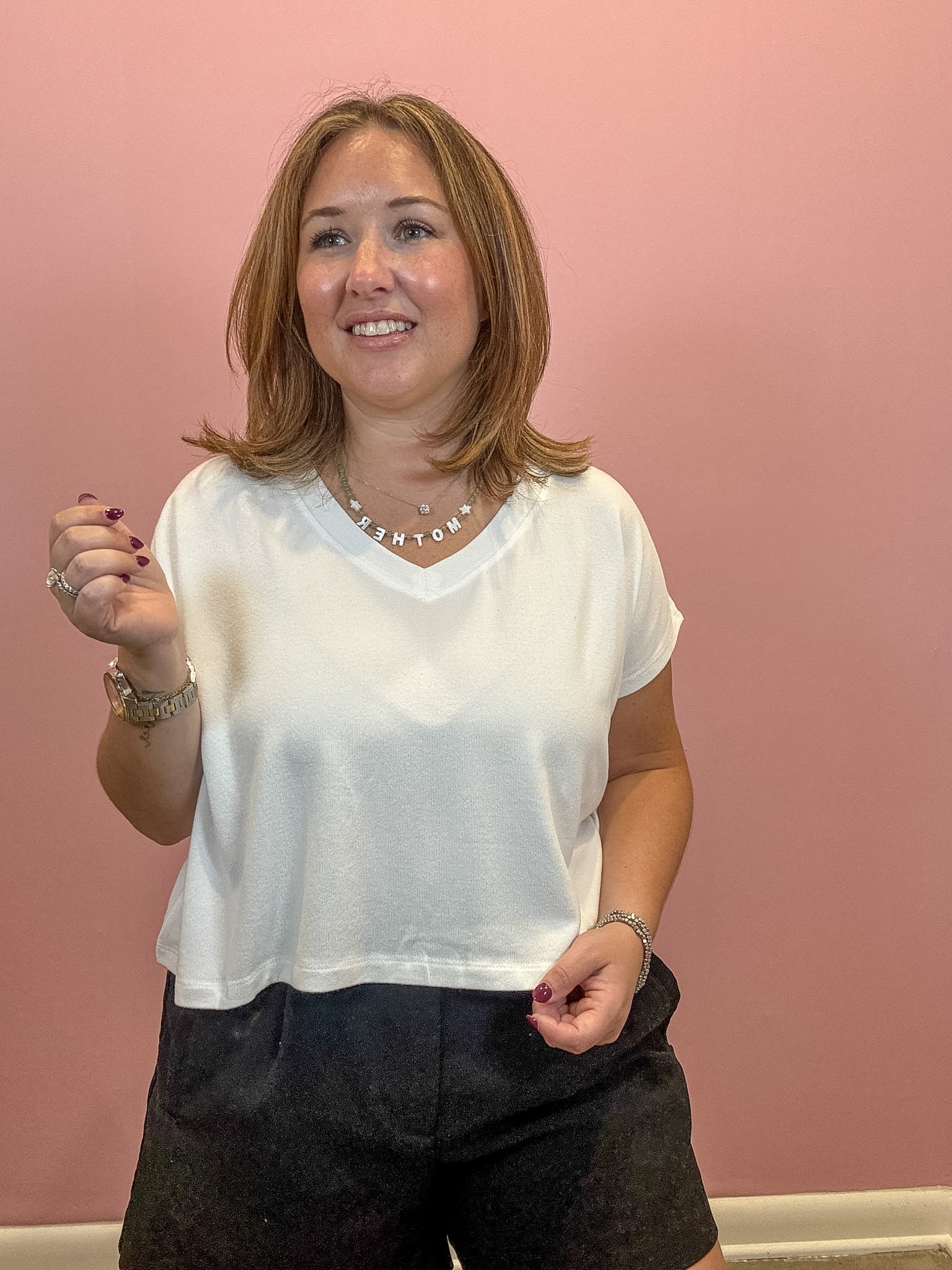 A woman with shoulder-length brown hair smiling while wearing a white short-sleeve V-neck top and dark shorts, standing against a solid pink wall.