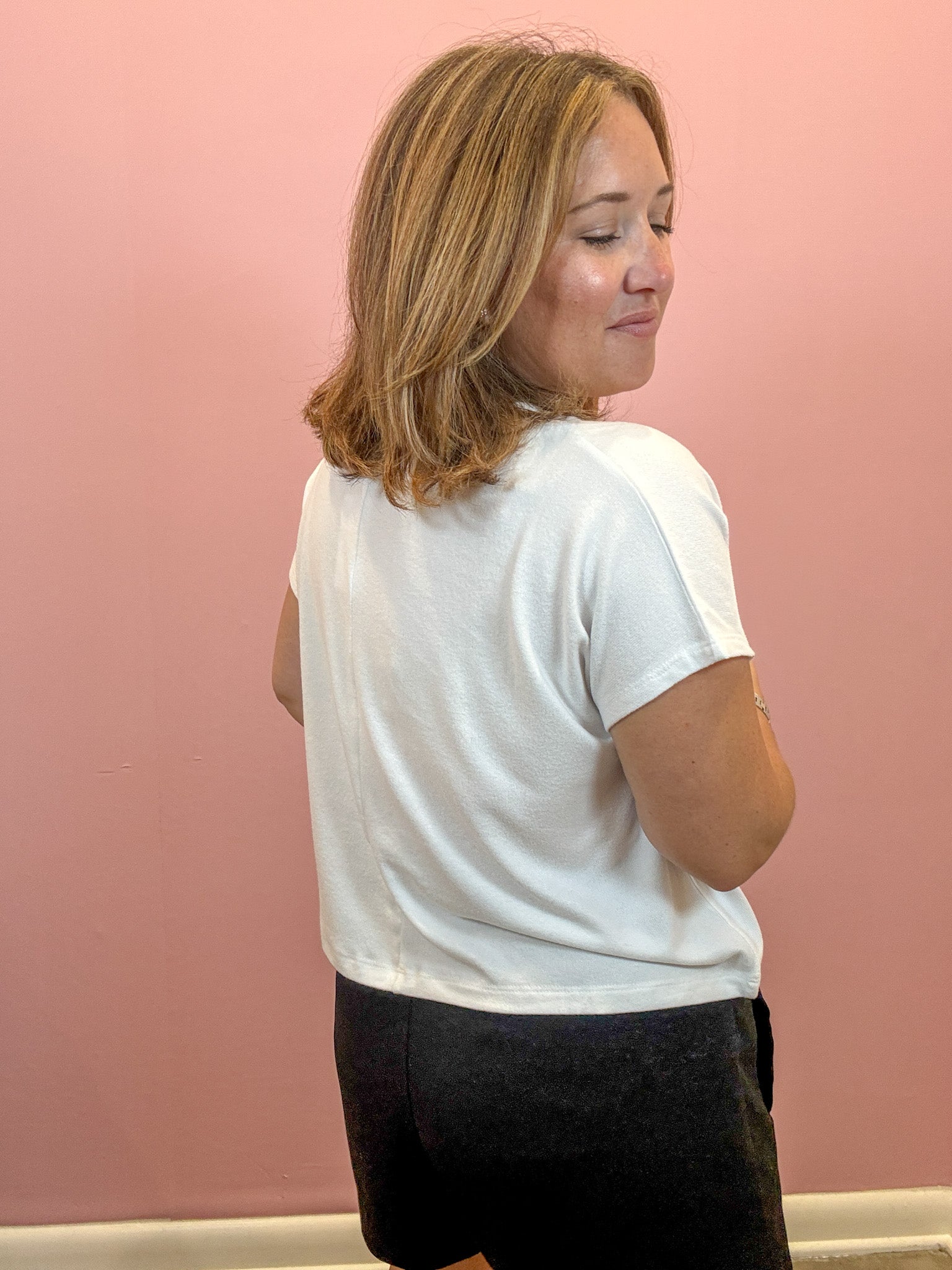 A woman with shoulder-length blonde hair is shown from the back wearing a white short-sleeve t-shirt and black shorts, standing against a solid pink wall.