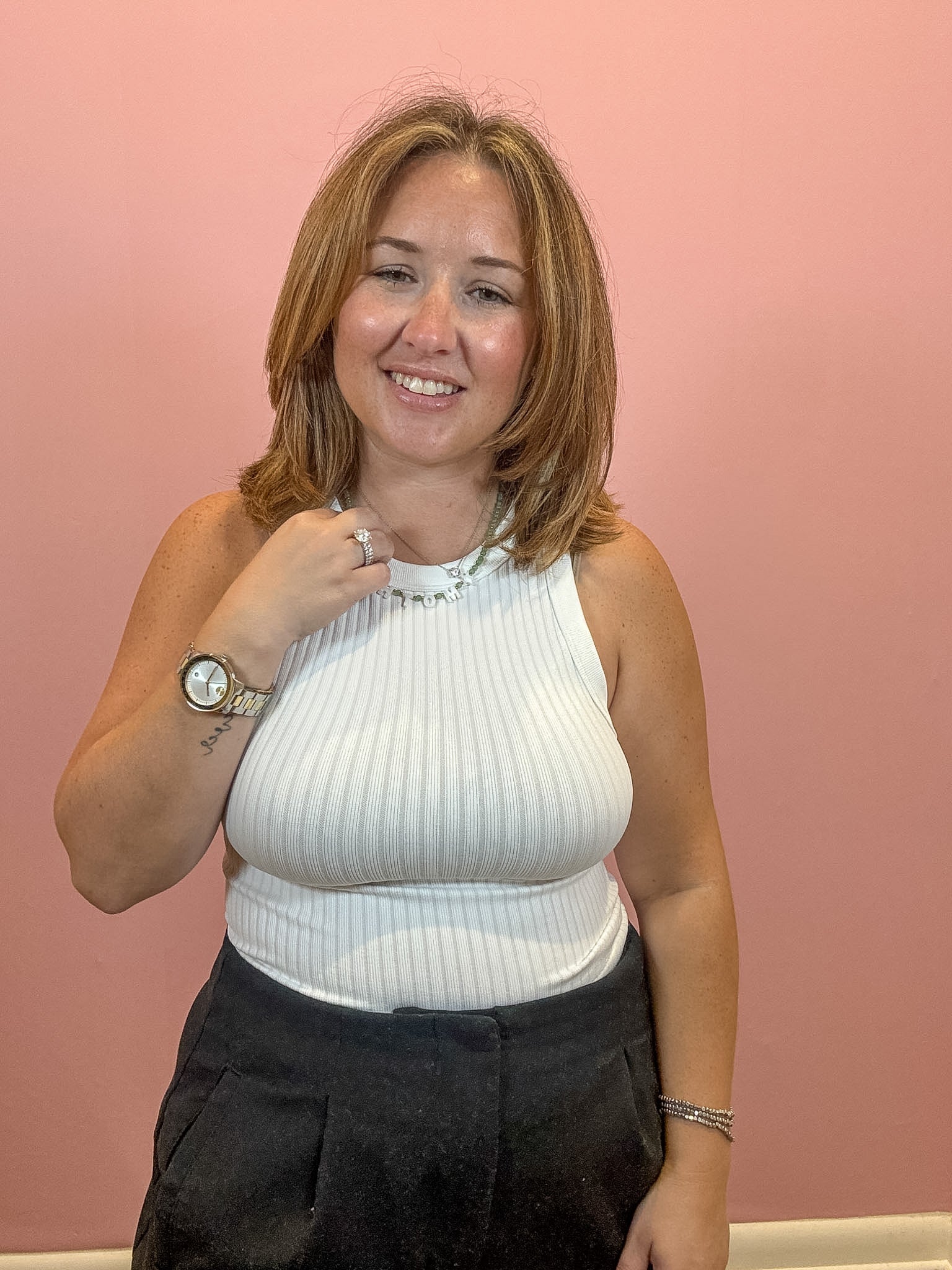 A woman with shoulder-length brown hair smiling in front of a solid pink wall, wearing a white ribbed halter top and dark grey trousers.