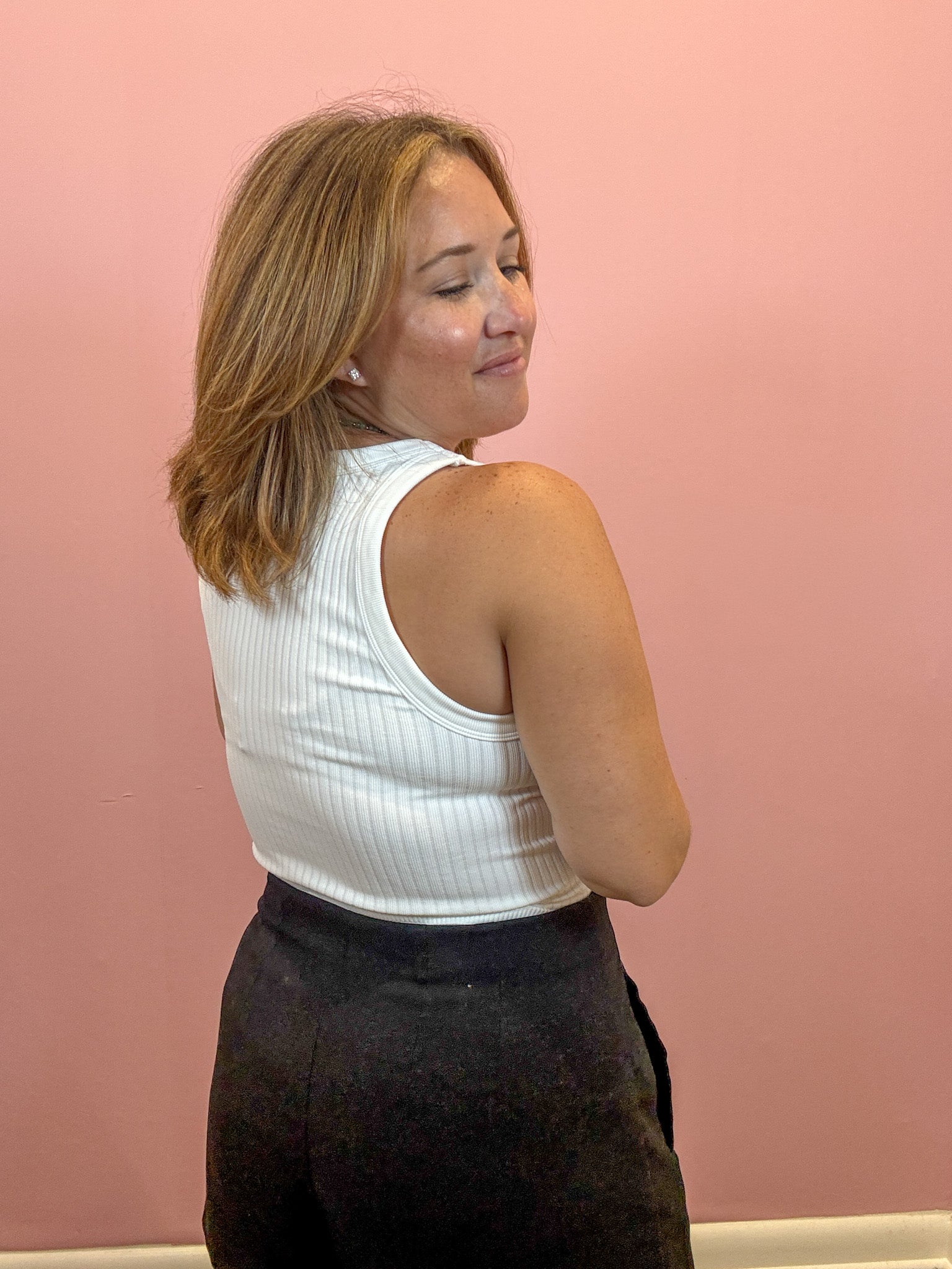 A woman with shoulder-length blonde hair is shown from the back, wearing a white ribbed tank top and dark charcoal pants, standing against a solid pink wall.
