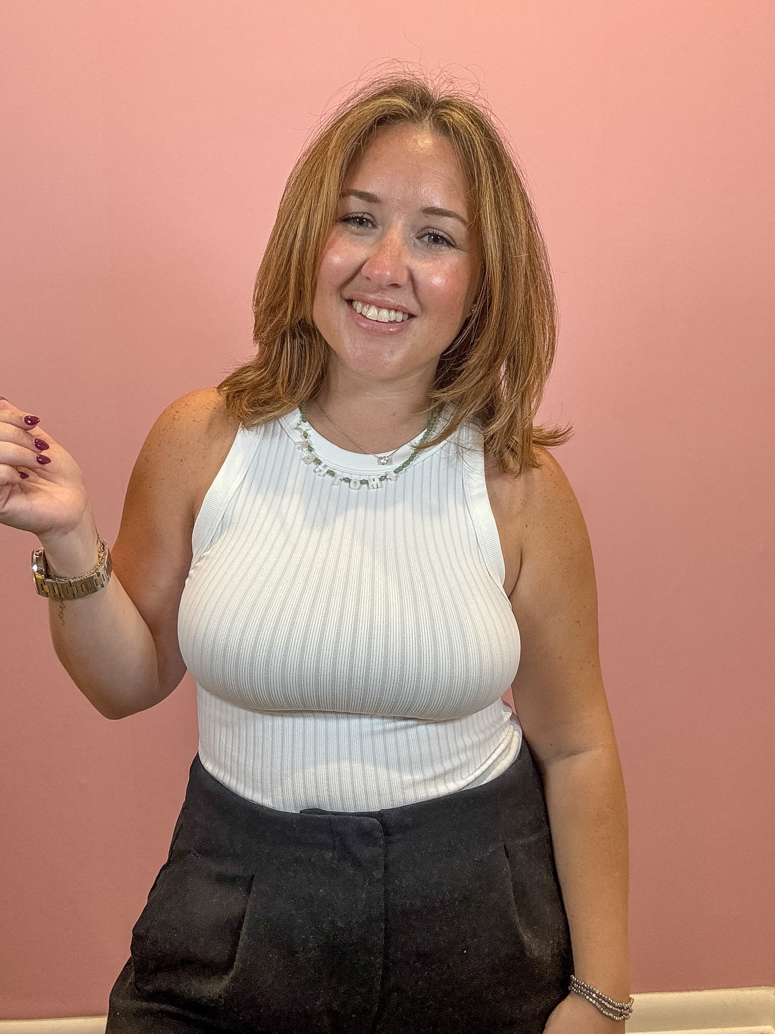 A smiling woman with shoulder-length light brown hair wearing a white ribbed halter top and dark pants, standing against a solid pink wall.