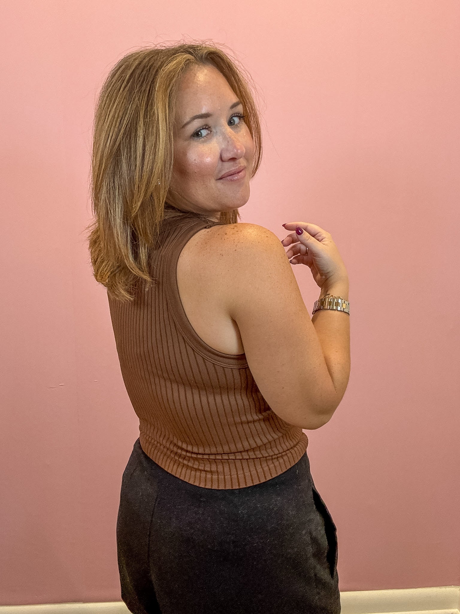 A woman with shoulder-length blonde hair is shown from the back wearing a brown ribbed sleeveless top and dark gray pants, posing against a solid pink wall.