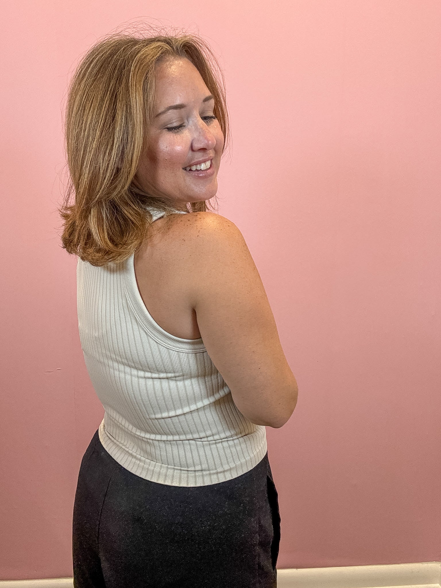 A woman with shoulder-length blonde hair is shown from the back, wearing a cream ribbed tank top and dark pants, standing against a solid pink wall.