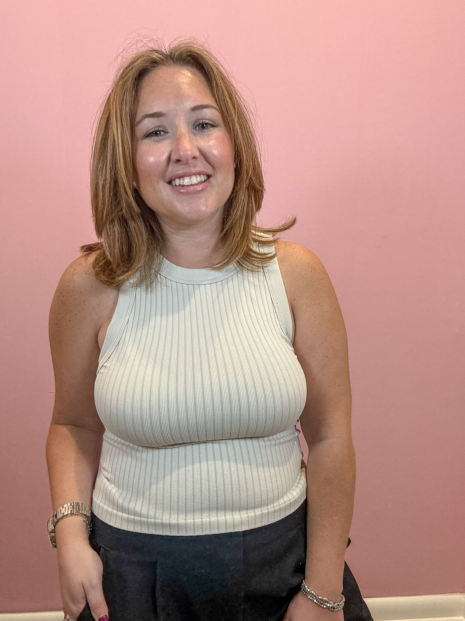 A smiling woman with shoulder-length blonde hair wearing a cream ribbed halter top and dark trousers, standing against a solid pink wall.