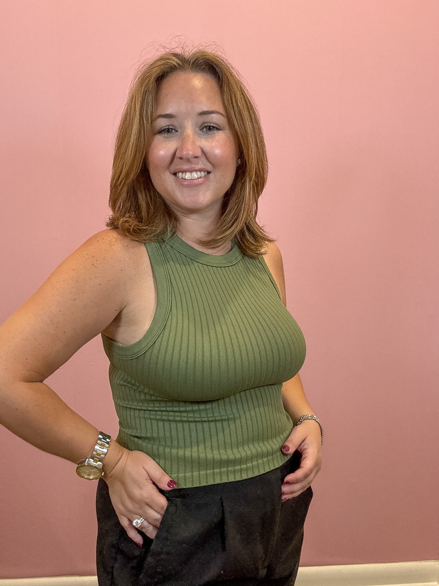 A smiling woman with shoulder-length light brown hair wearing a ribbed olive green sleeveless tank top and dark pants, standing against a solid pink wall.