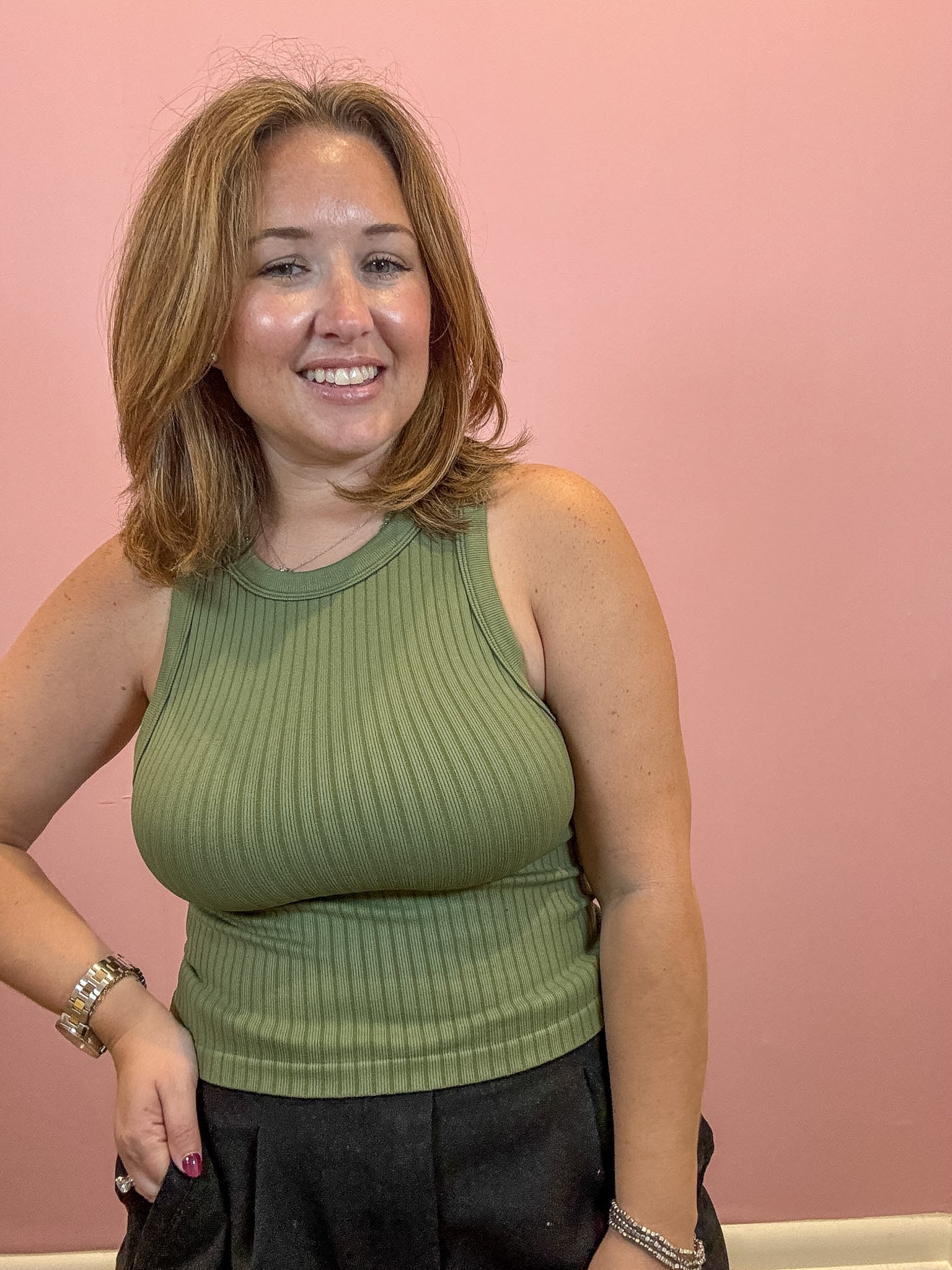 A smiling woman with shoulder-length blonde hair wearing a ribbed olive green sleeveless tank top and black pants, standing against a solid pink wall.