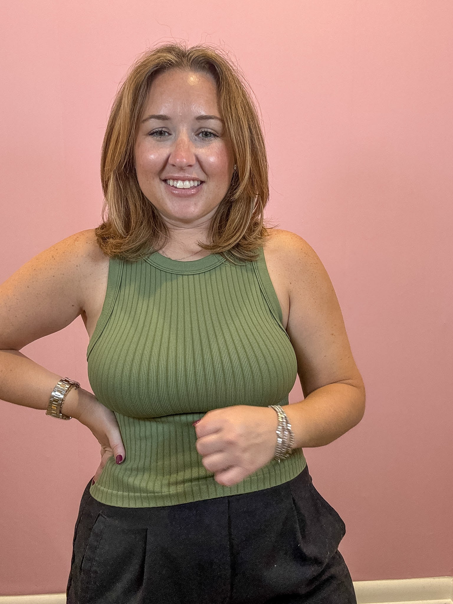 A smiling woman with shoulder-length light brown hair wearing a ribbed olive green sleeveless tank top and dark pants, standing against a solid pink wall.