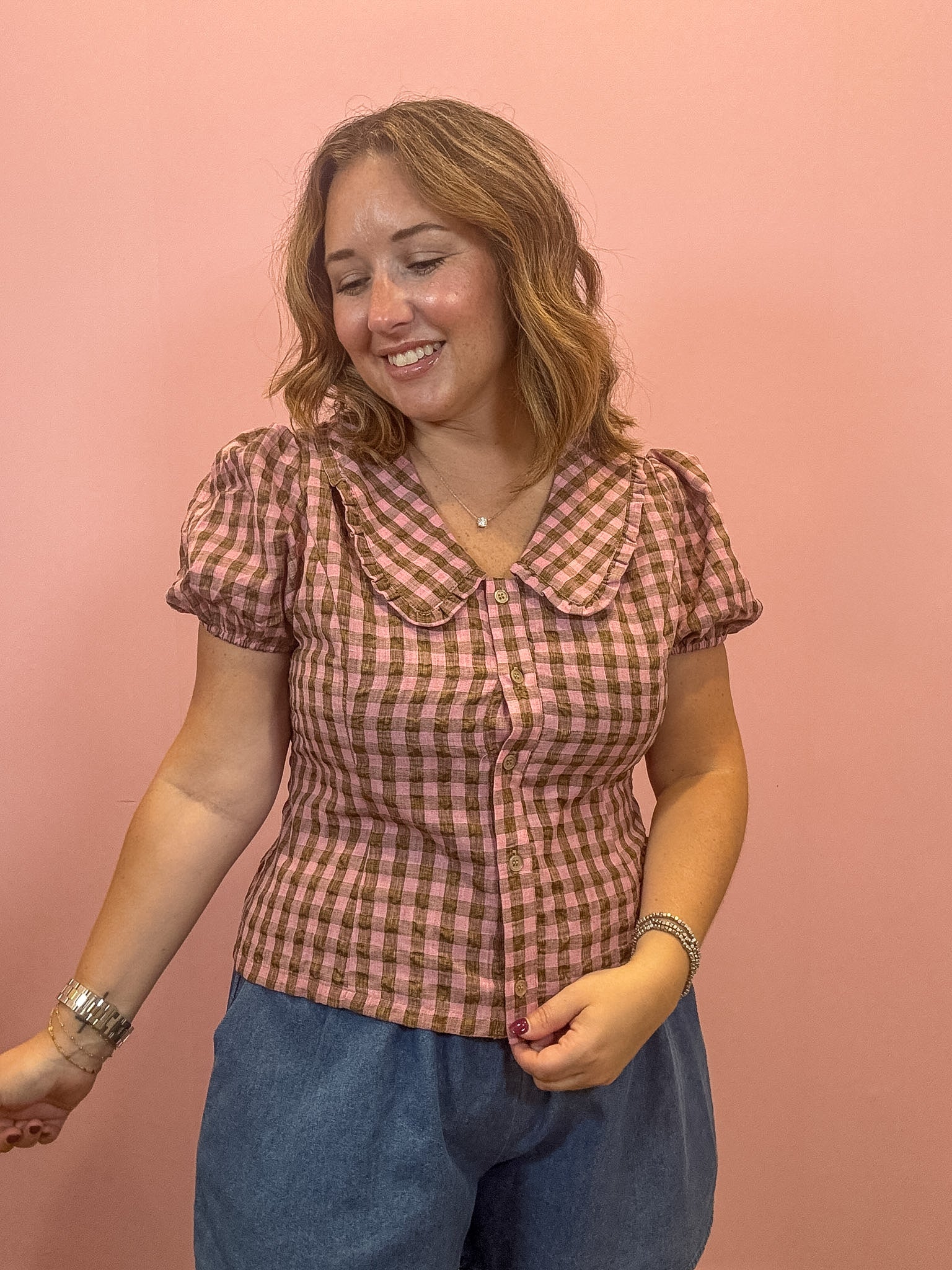 A woman wearing a pink and brown gingham short-sleeve blouse with a Peter Pan collar, paired with blue jeans, standing against a solid pink background.