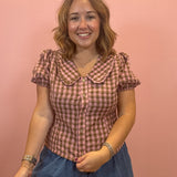 A woman modeling a pink and brown gingham short-sleeve blouse with a Peter Pan collar, paired with blue denim shorts, standing against a solid pink studio backdrop.