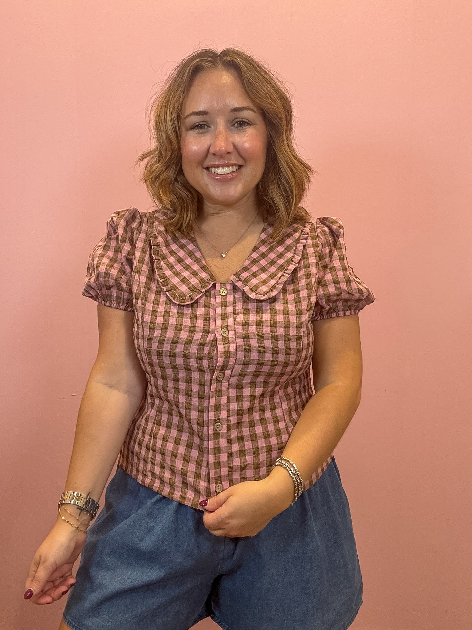 A woman modeling a pink and brown gingham short-sleeve blouse with a Peter Pan collar, paired with blue denim shorts, standing against a solid pink studio backdrop.