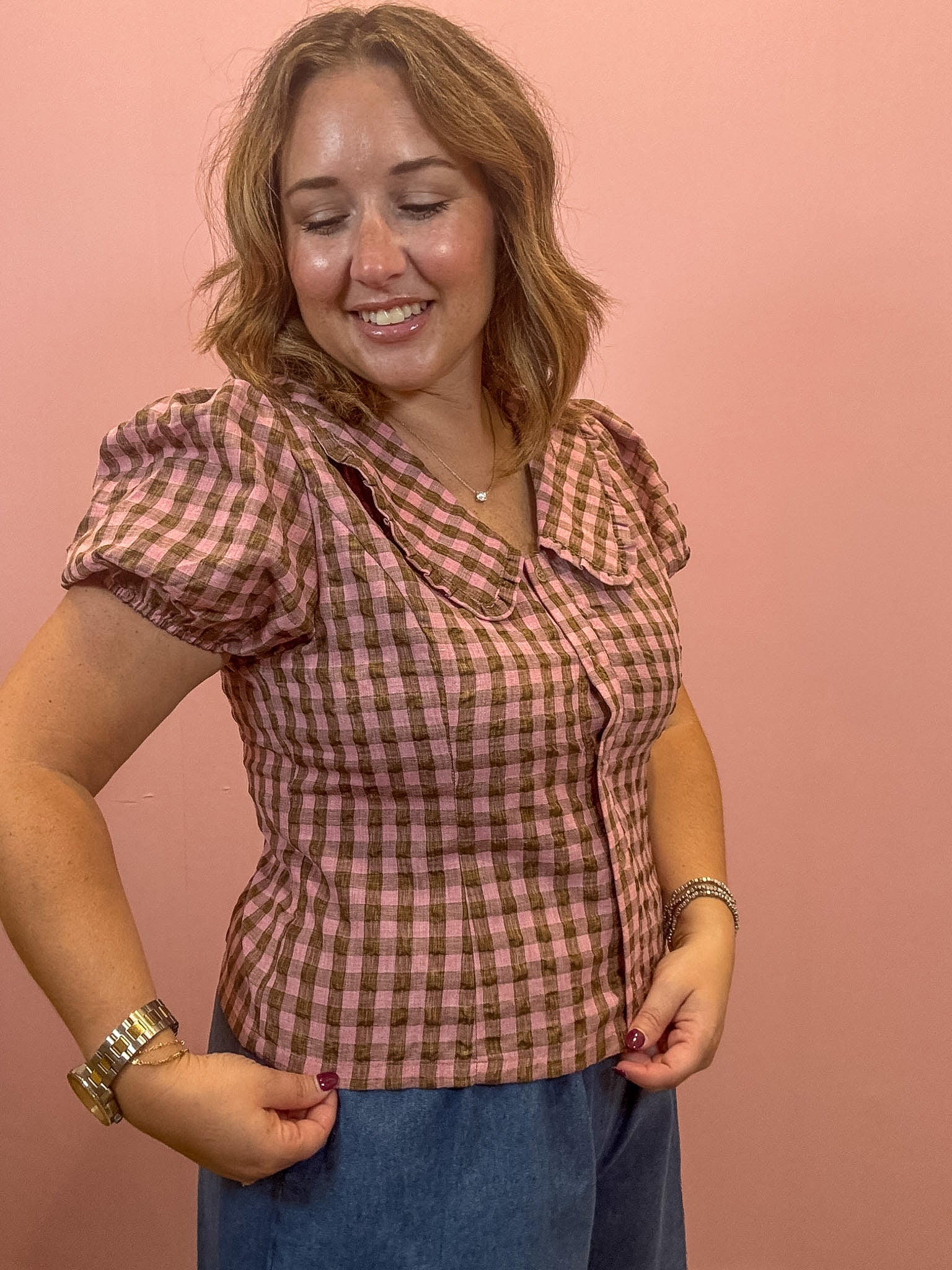 A woman wearing a pink and brown gingham short-sleeve blouse with puff sleeves and a Peter Pan collar, paired with blue jeans, standing against a solid pink background.