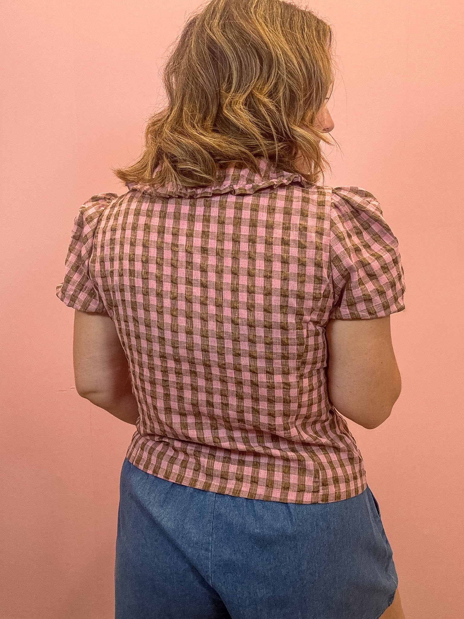 A woman viewed from behind wearing a pink and brown gingham short-sleeve blouse with puff sleeves and blue denim shorts, standing against a solid pink background.