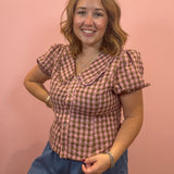 A woman wearing a pink and brown gingham short-sleeve button-up shirt with puff sleeves and a Peter Pan collar, paired with blue denim shorts, posing against a solid pink wall.